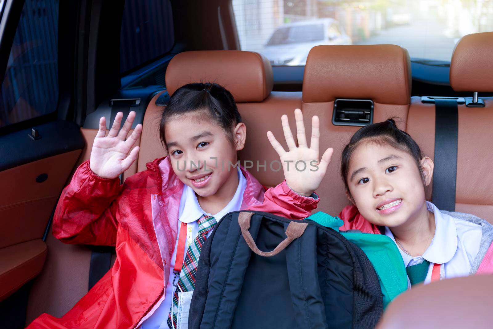 Two happy students raising hands and motion while enjoying road trip in car. Sisters smiling and looking camera inside car in morning. Asian cildrens sitting in car on the way go to school. Family in car
