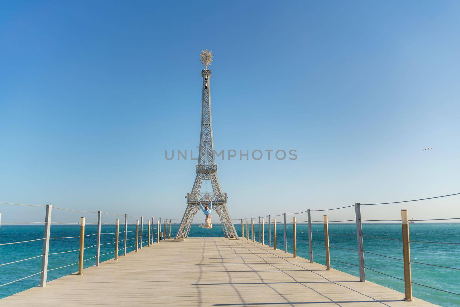 Large model of the Eiffel Tower on the beach. A woman walks along the pier towards the tower, wearing a blue jacket and white jeans. by Matiunina