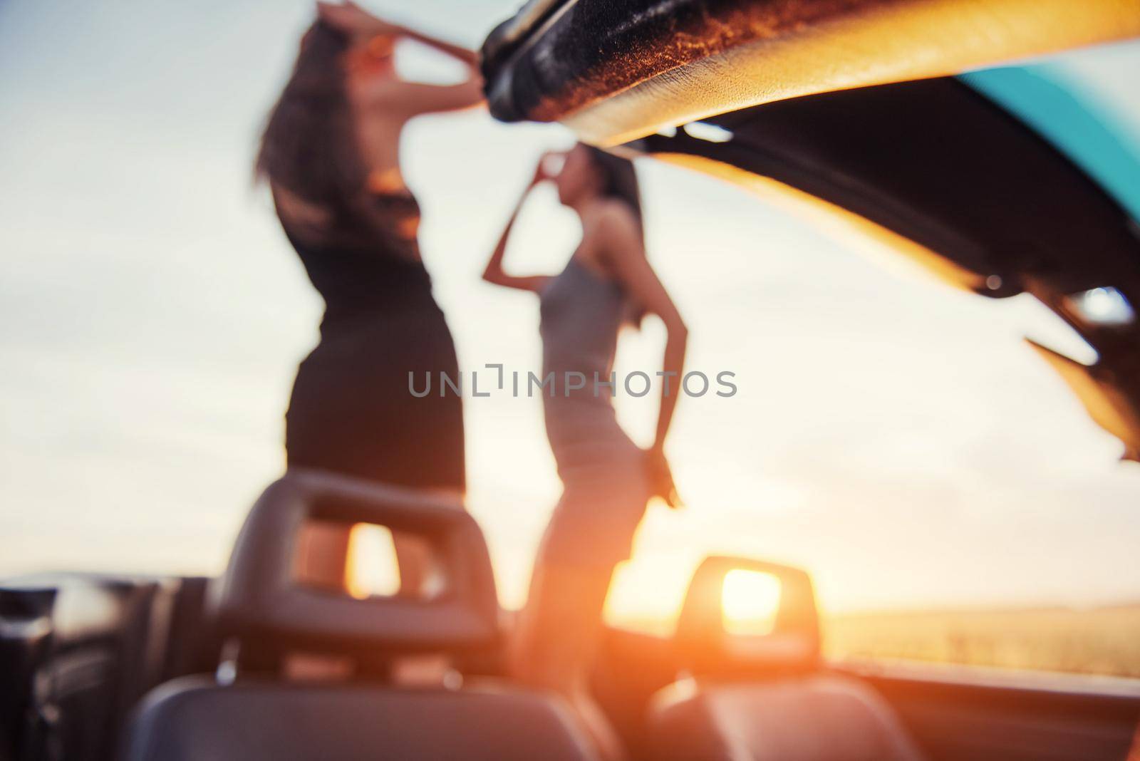 Two girls happy to pose next to a black car against the sky on a fantastic sunset. Natural blurred background. Soft light effect.