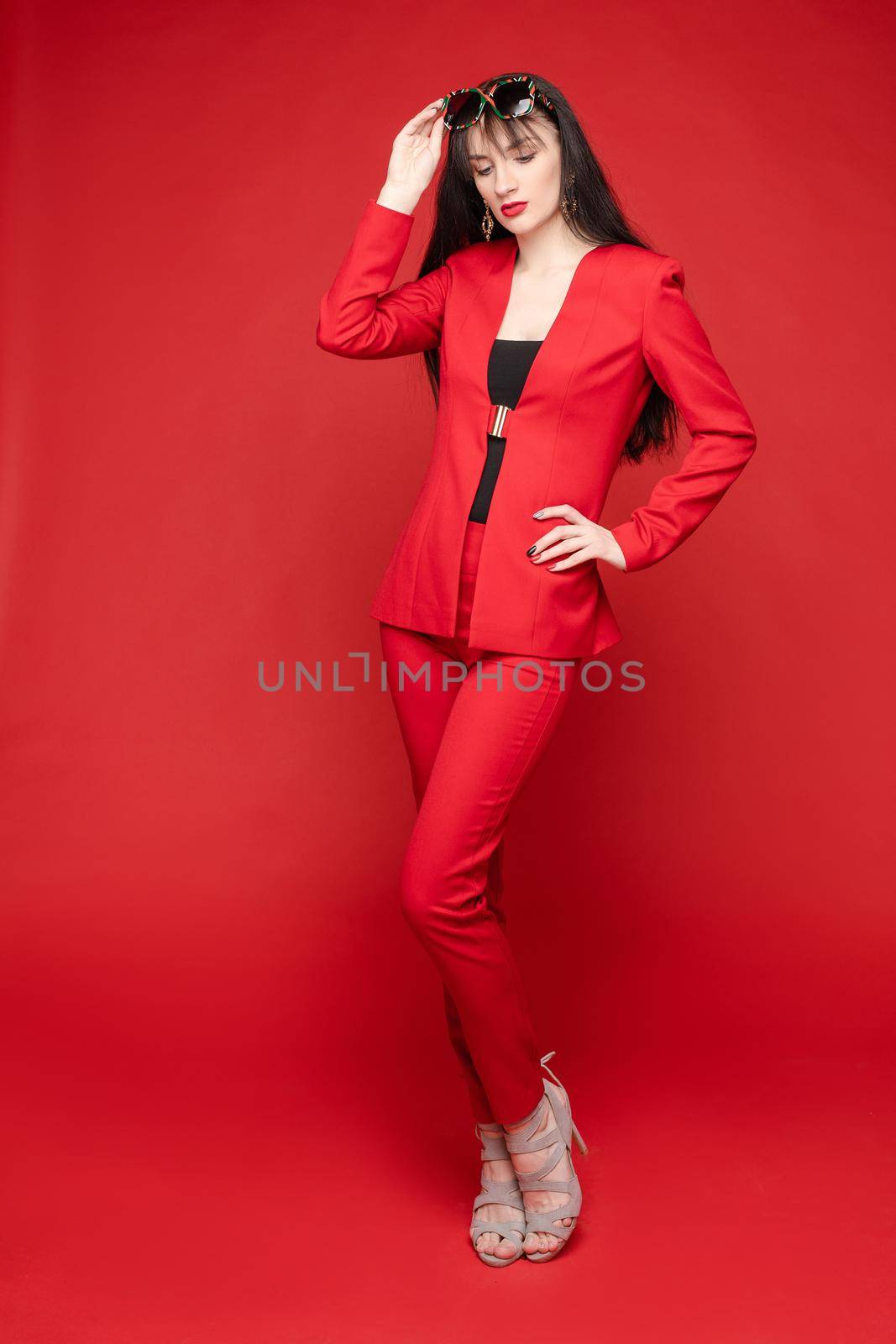Side view of glamorous brunette posing in red smart suit and heels on red isolated background in studio. Young woman keeping glasses in hand and looking down. Concept of beauty and fashion.