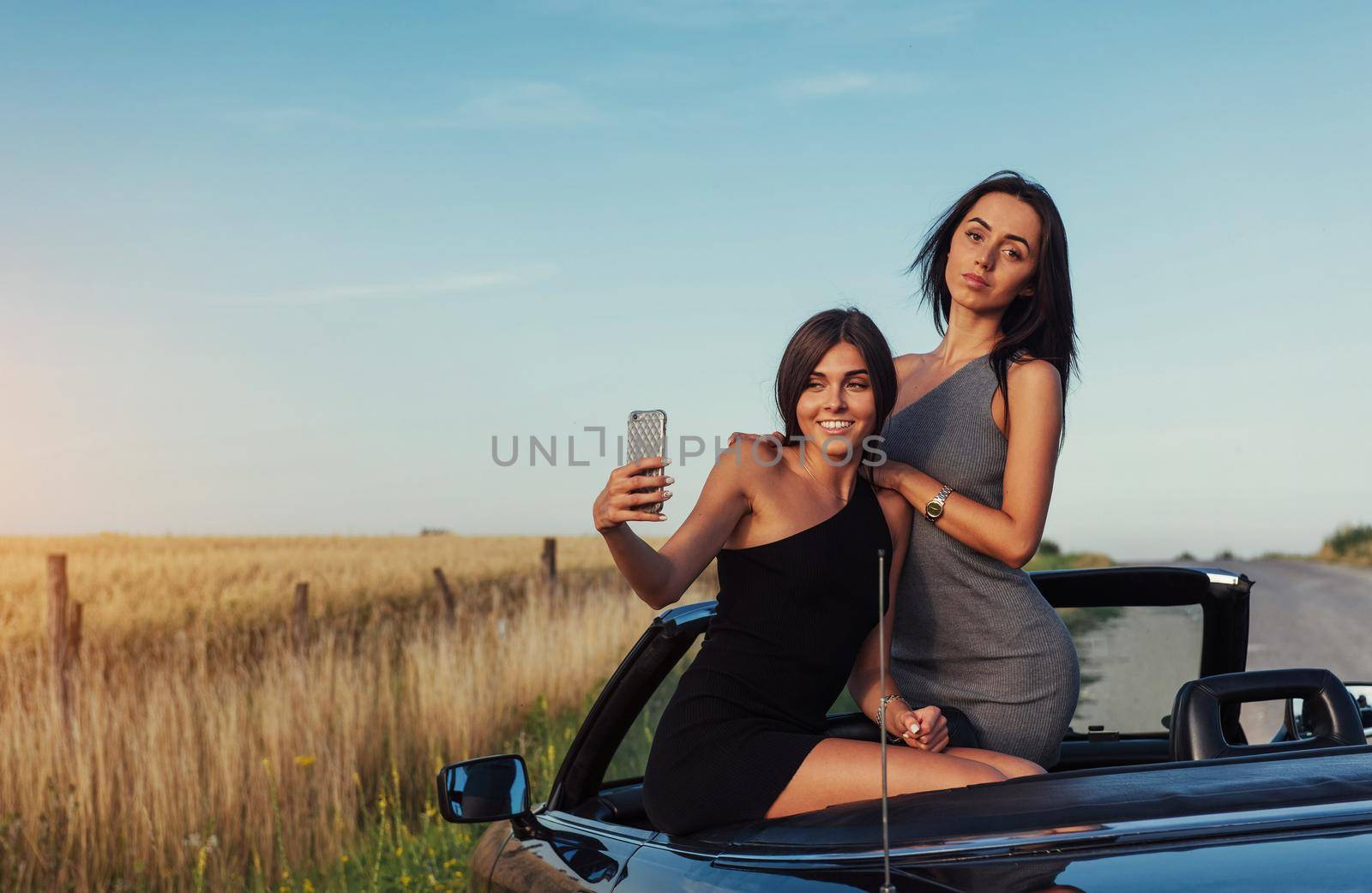 Beautiful two girls are photographed on the road against a background of blue sky and field on a black convertible.