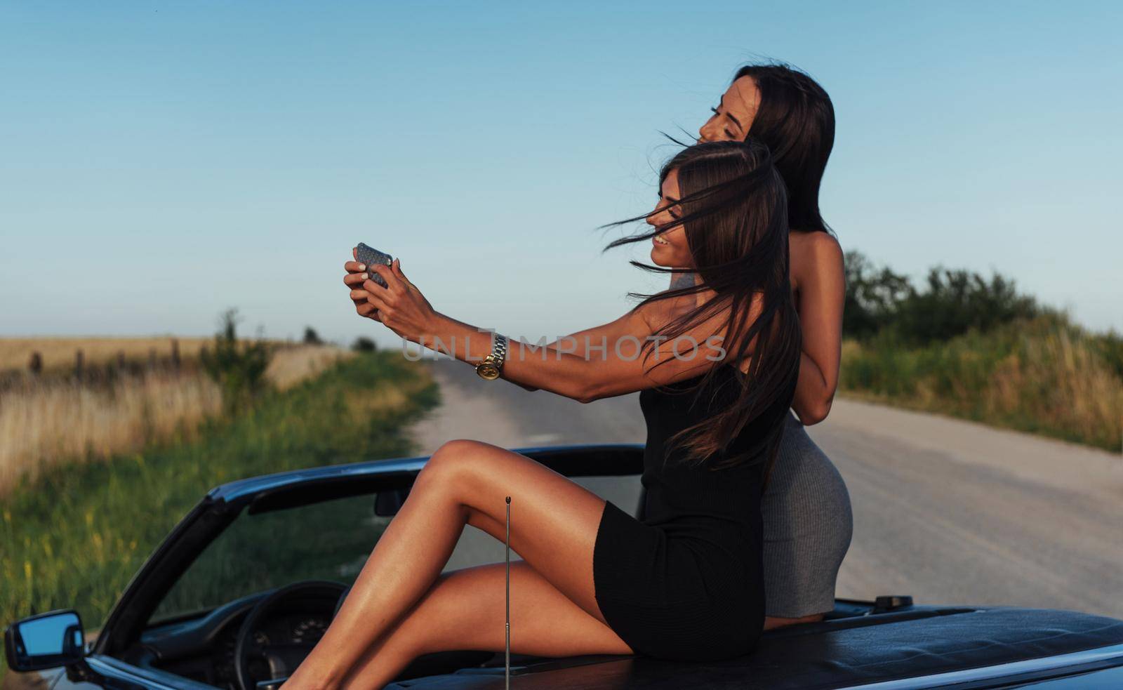 Beautiful two girls are photographed on the road against a background of blue sky and field on a black convertible.