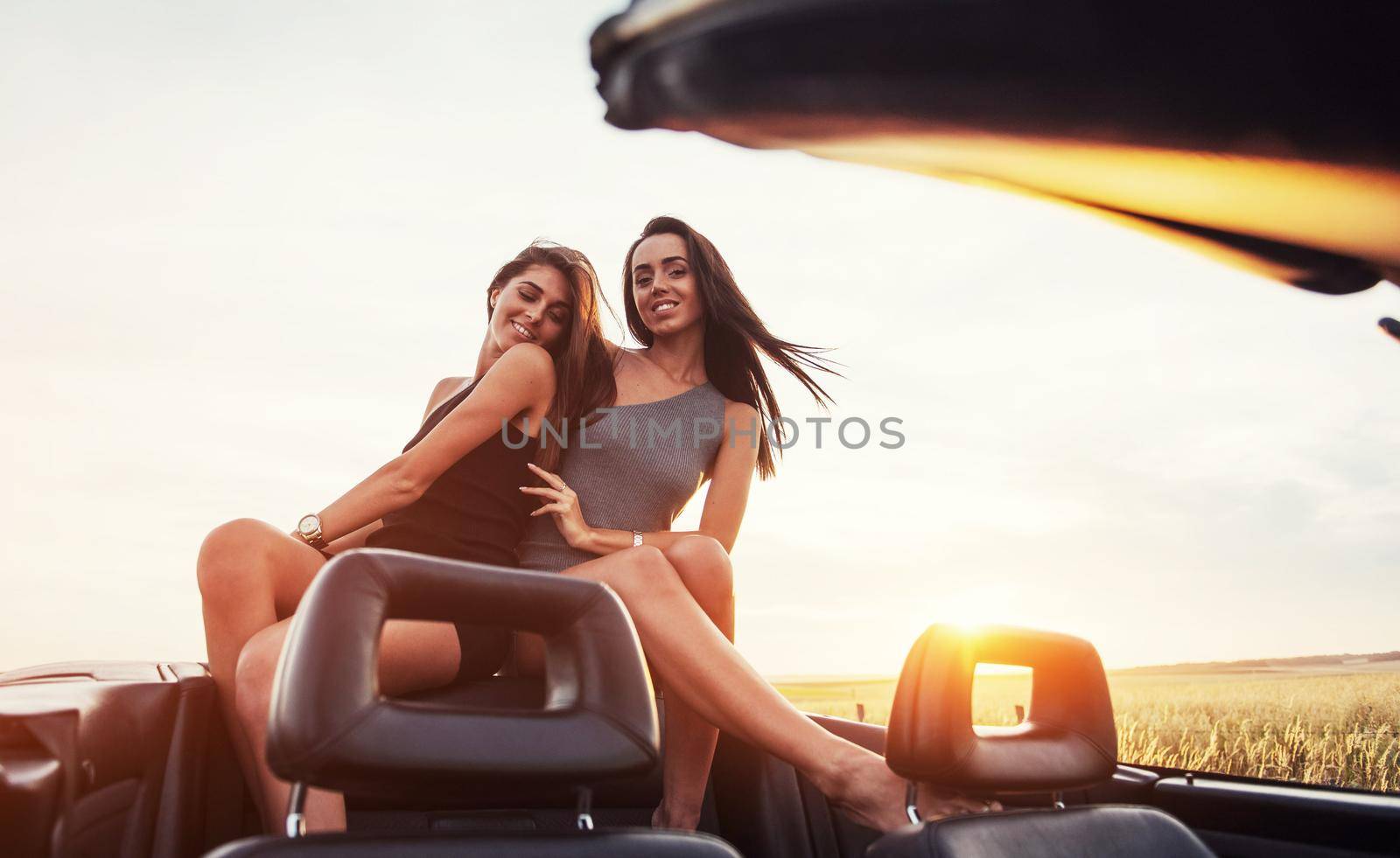 Young two women at a photo shoot. Girls gladly posing next to a black car against the sky on a fantastic sunset.