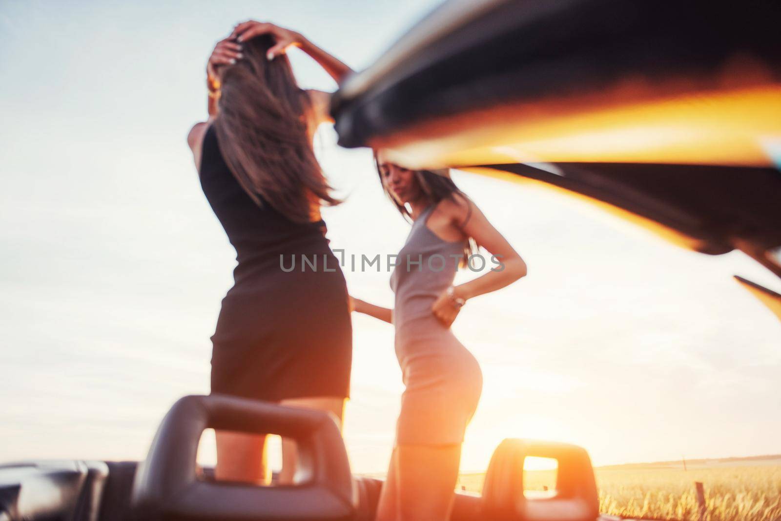Girls gladly posing next to a black car against the sky on a fantastic sunset.