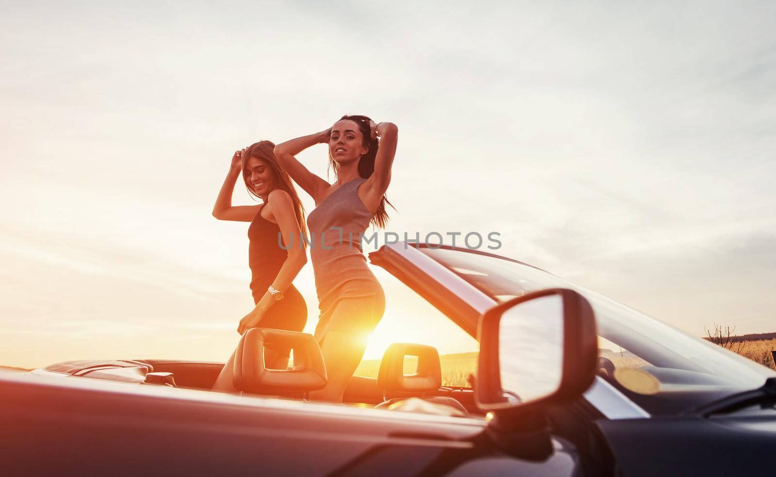 Young two women at a photo shoot. Girls gladly posing next to a black car against the sky on a fantastic sunset.