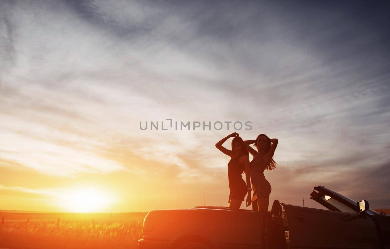 Young two women at a photo shoot. Girls gladly posing next to a black car against the sky on a fantastic sunset.