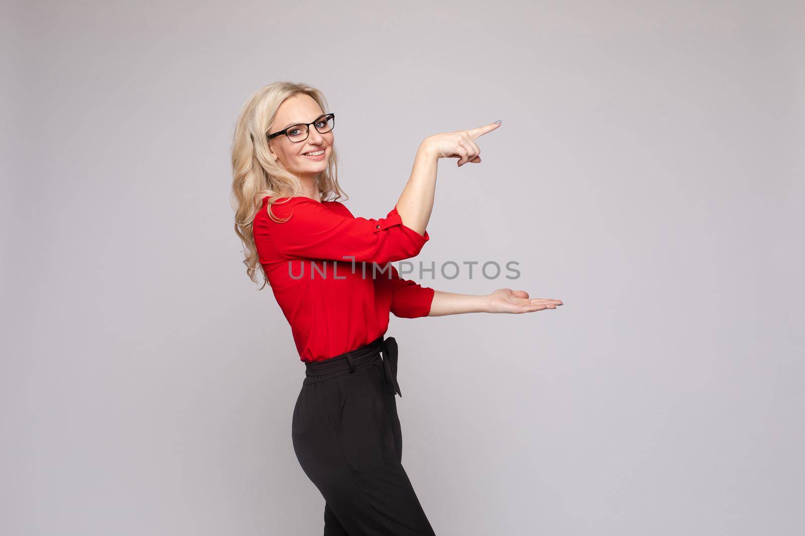 Side view of cheerful blonde in glasses, black skirt and red blouse looking at camera and gesturing with hands. Attractive woman keeping in hands copy space for text and object on isolated background.