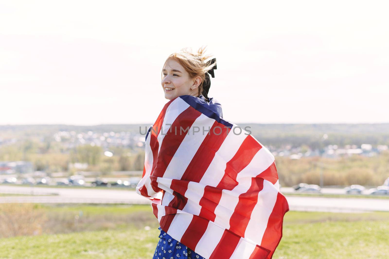 A cute young woman is wrapped in a usa flag and looks out over the city. 4th of July.