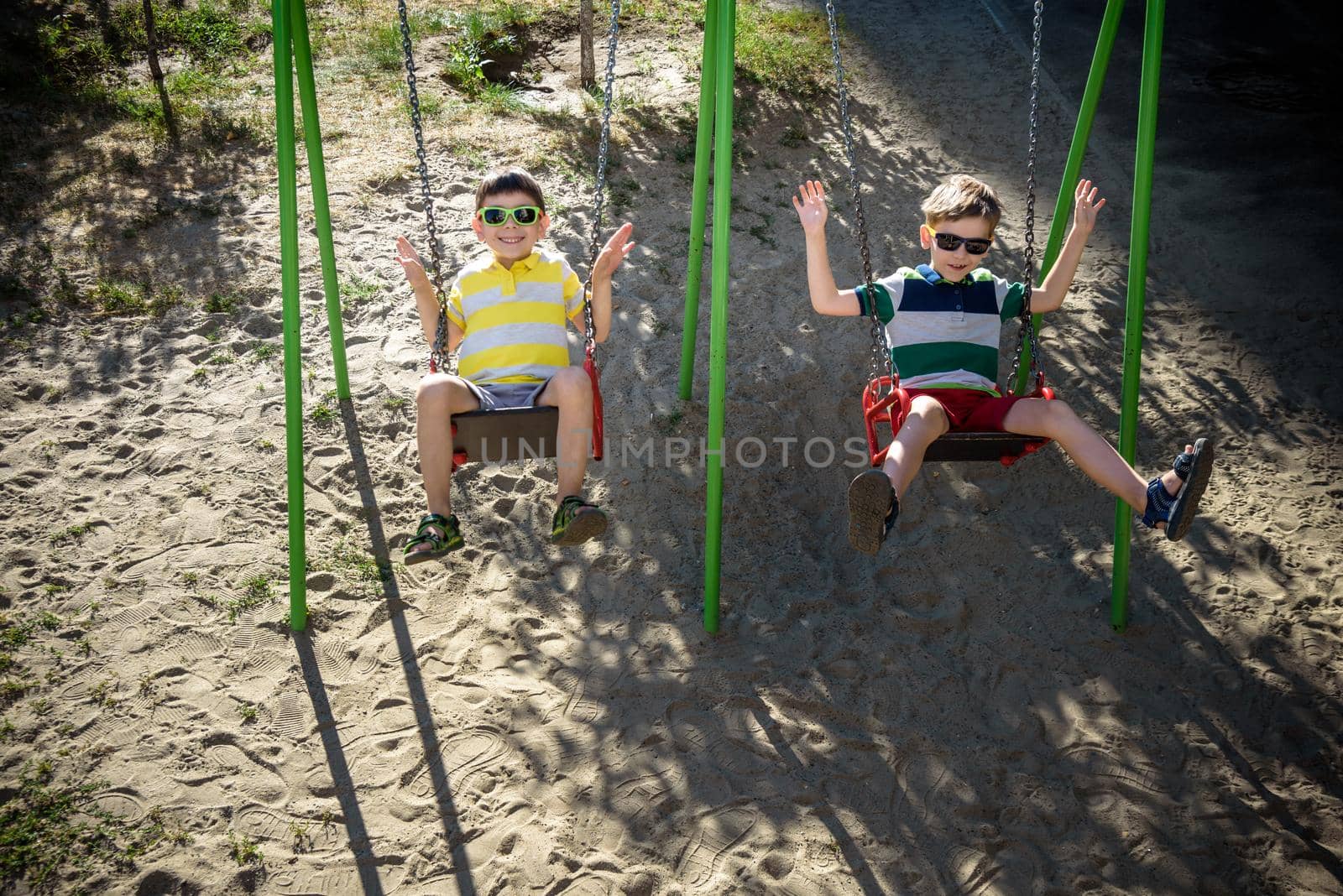 Two little kid boys having fun with swing on outdoor playground. Children, best friends and siblings swinging on warm sunny spring or autumn day. Active leisure with kids. Casual boy fashion.