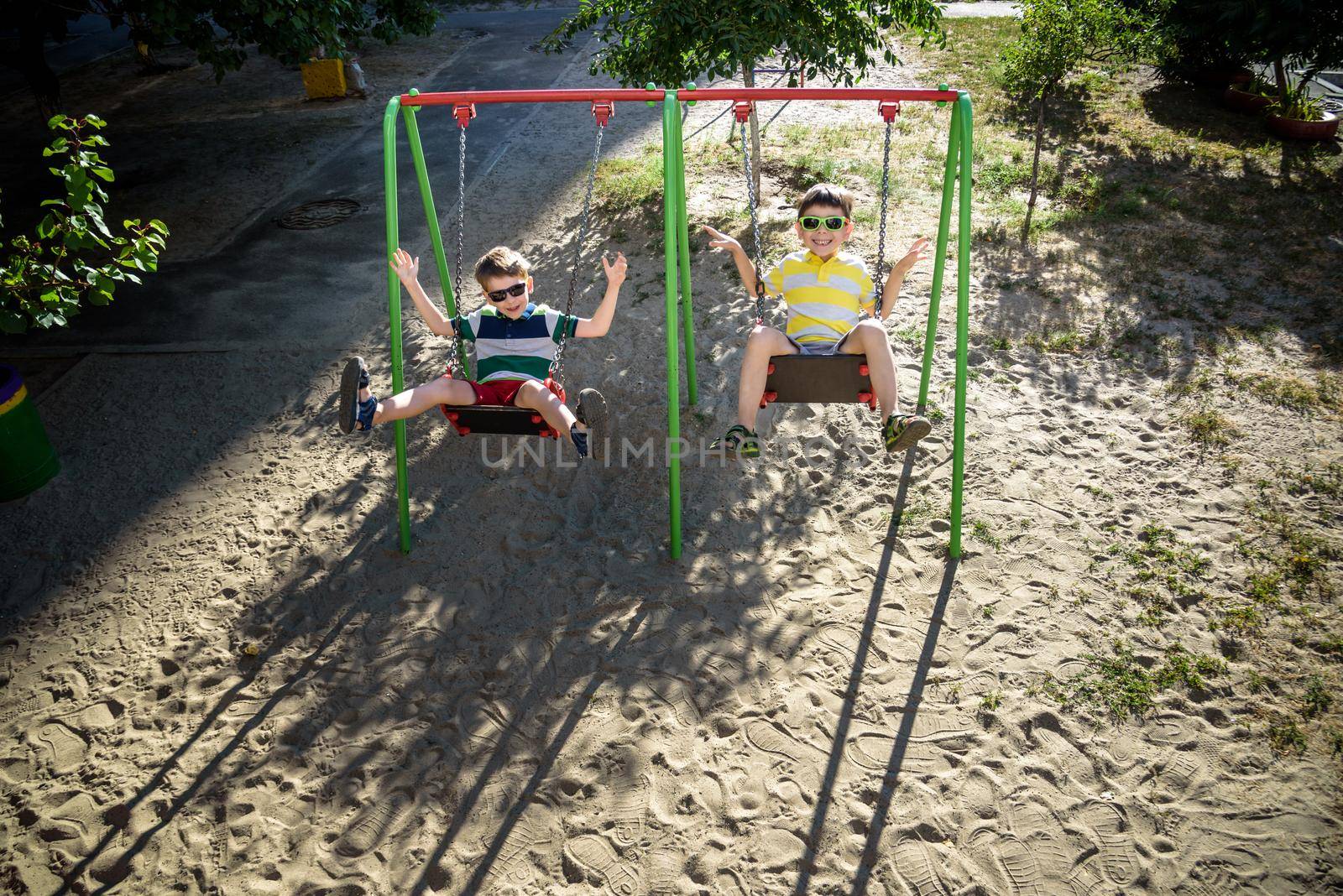 Two little kid boys having fun with swing on outdoor playground. Children, best friends and siblings swinging on warm sunny spring or autumn day. Active leisure with kids. Casual boy fashion.