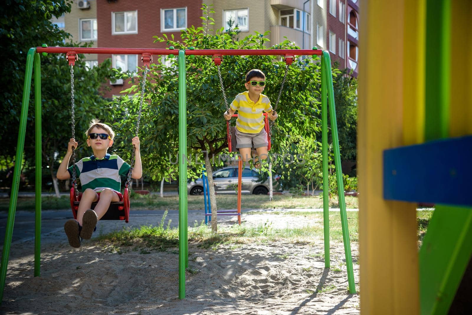 Two little kid boys having fun with swing on outdoor playground. Children, best friends and siblings swinging on warm sunny spring or autumn day. Active leisure with kids. Casual boy fashion.