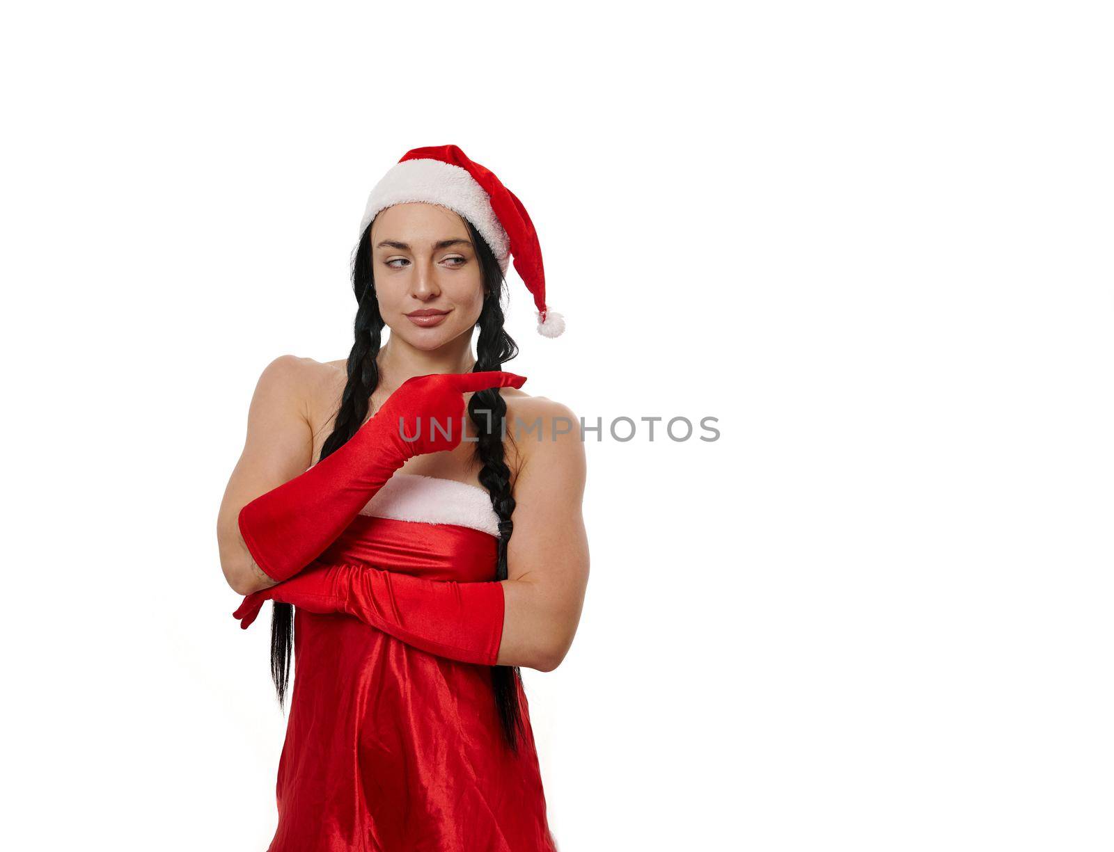 Beautiful sexy dark-haired woman in a carnival costume of Santa Claus smiling cute, looking mysteriously aside and pointing with finger on a copy space of a white wall background. Christmas concept