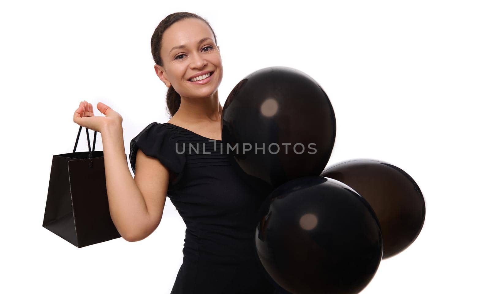 Black Friday concept with attractive mixed race woman with beautiful toothy smile, dressed in black looks at the camera stands with black air balloons and shopping bag, white background, copy space.