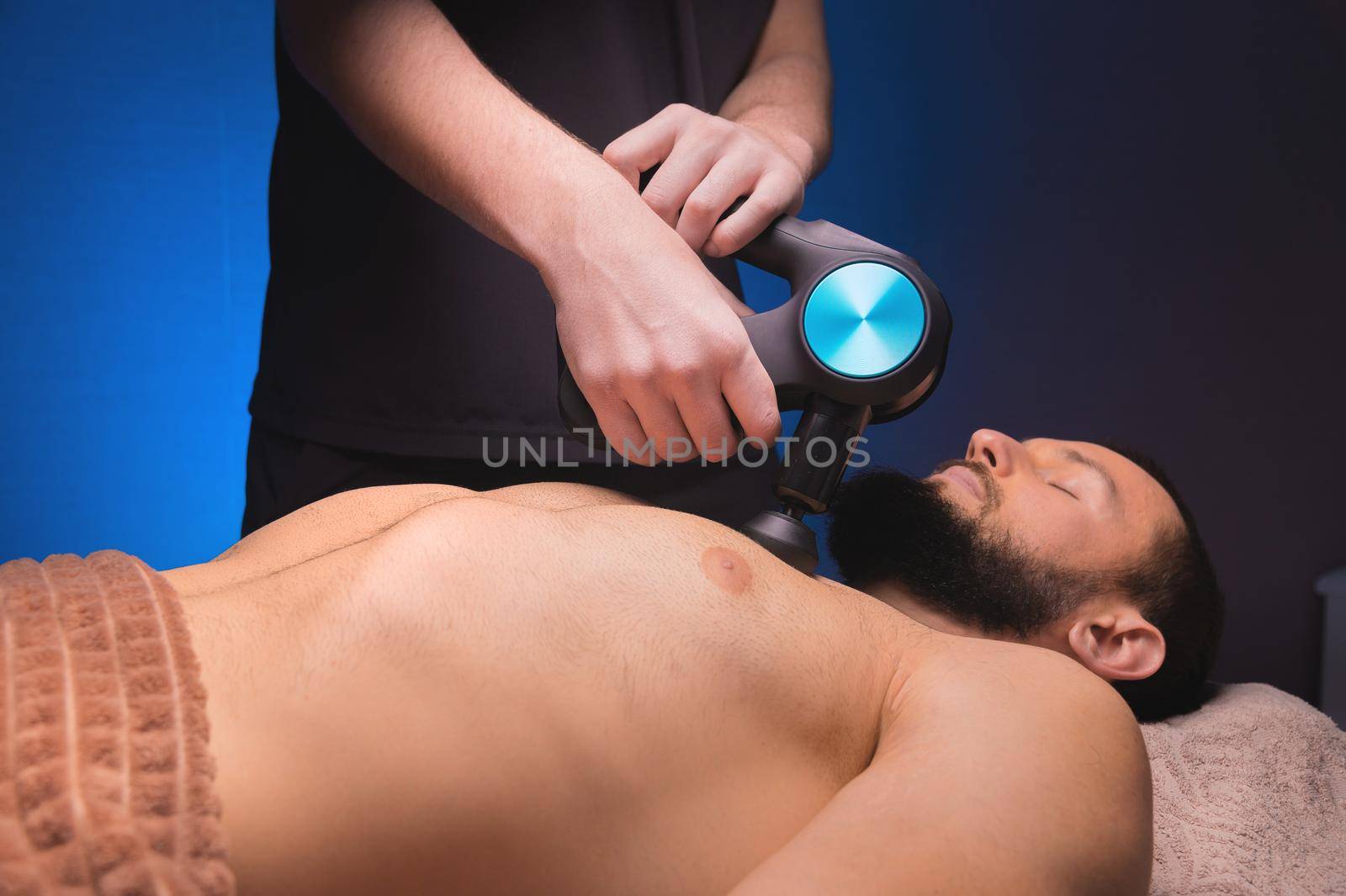 Professional massage of the chest and pectoral muscles with a percussion shock wave massager in a professional office with subdued light.