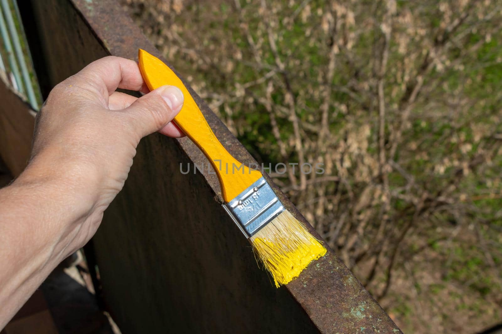 A hand with a yellow brush paints the rusty metal railing on the balcony on a sunny day. Diy.