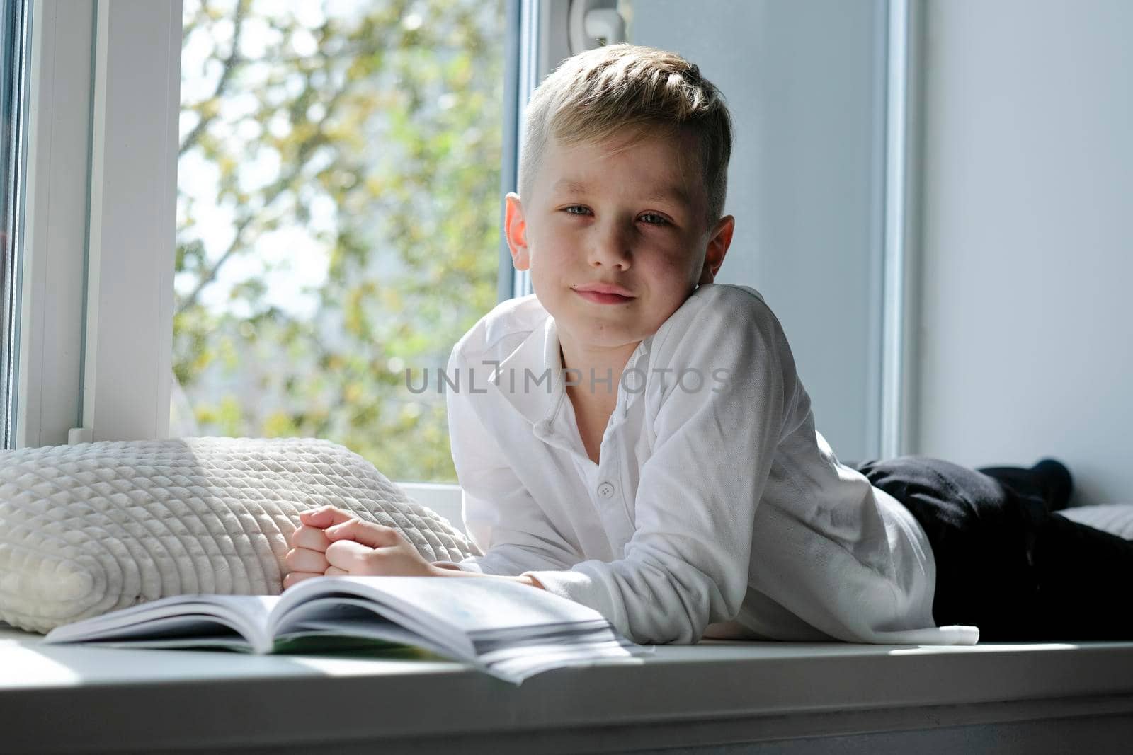 Cute boy reading a book lying on a windowsill in the house enjoying sunny weather
