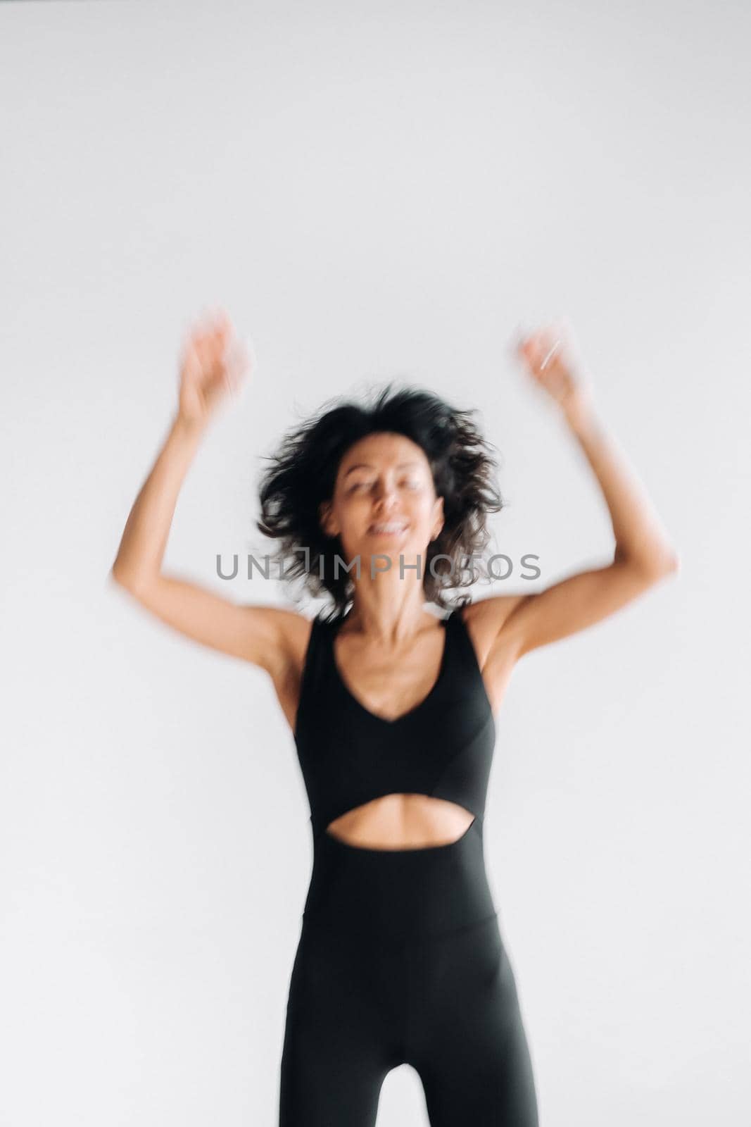 A blurry silhouette of a woman in black sportswear jumps up and raises her arms up against a white background.