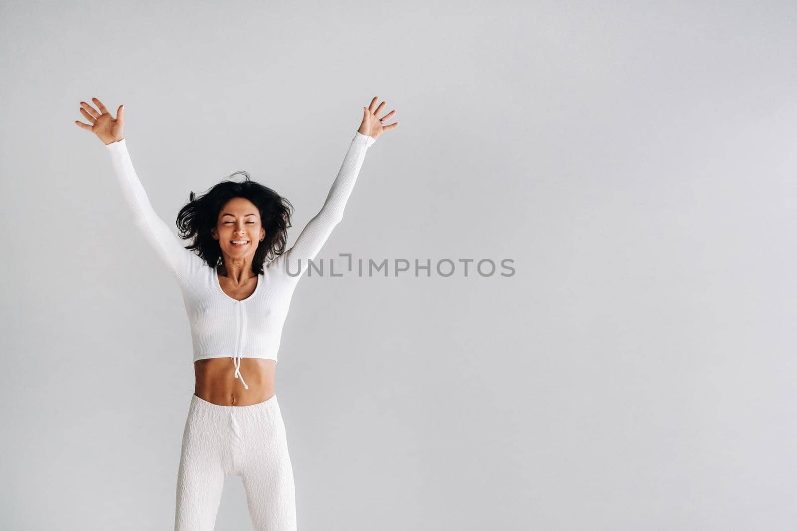 A happy woman in white sportswear bounces on a white background. The girl, jumping, raised her hands up in the gym.