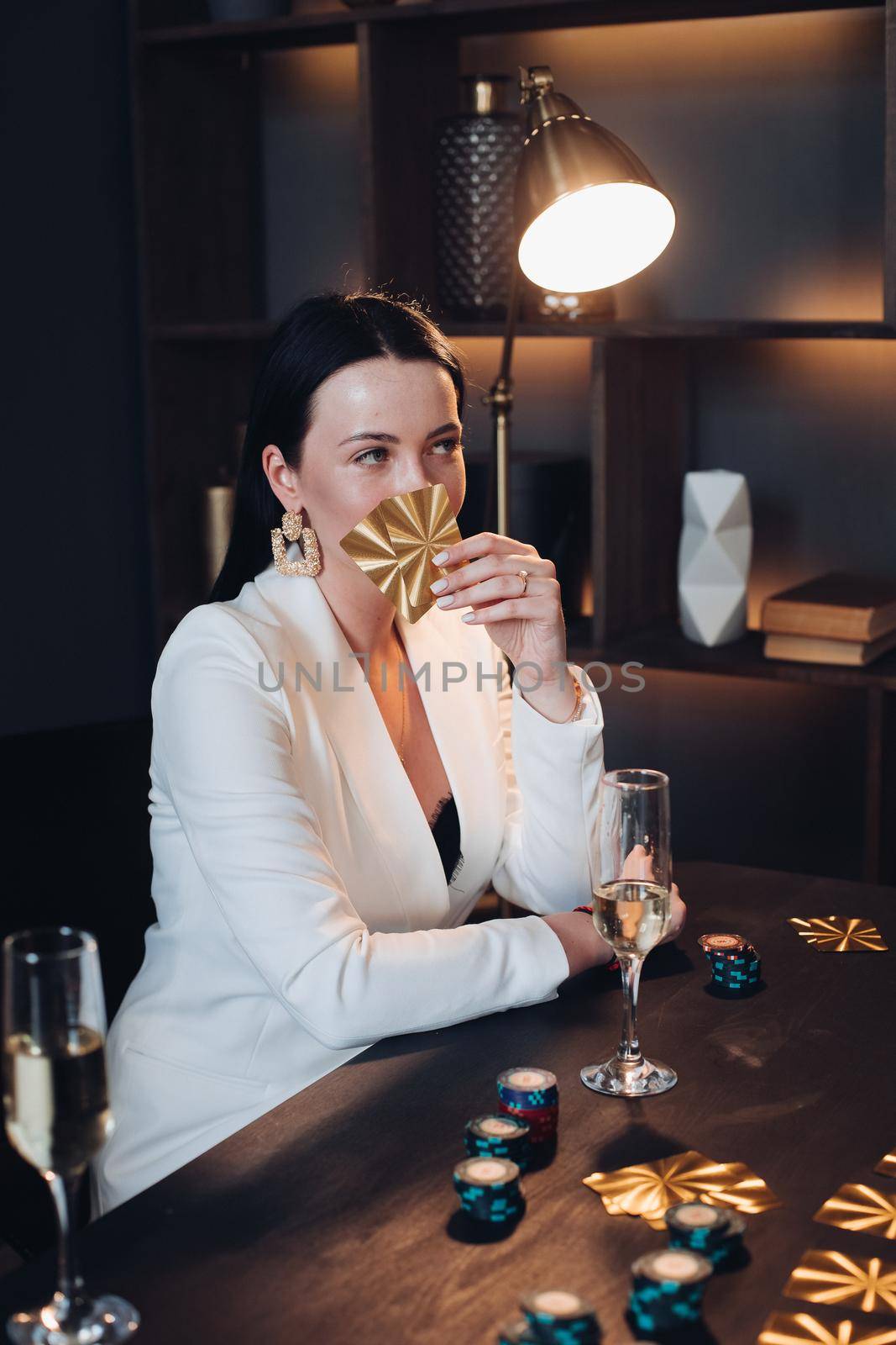 Stock photo portrait of gorgeous mystic woman in white jacket with black hair sitting at poker table with chips holding two cards in her hand and looking at her opponent. She is having a glass of champagne in front of her.