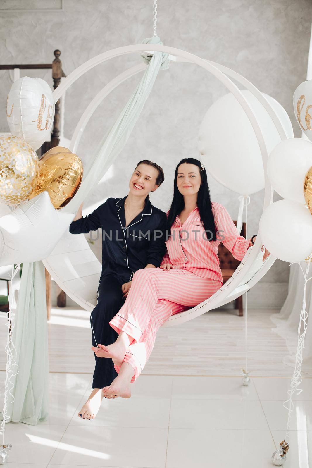 Two happy female friend in pajamas sitting on stylish armchair at luxury interior. Smiling girlfriends enjoying friendship surrounded by festive party event having positive emotion