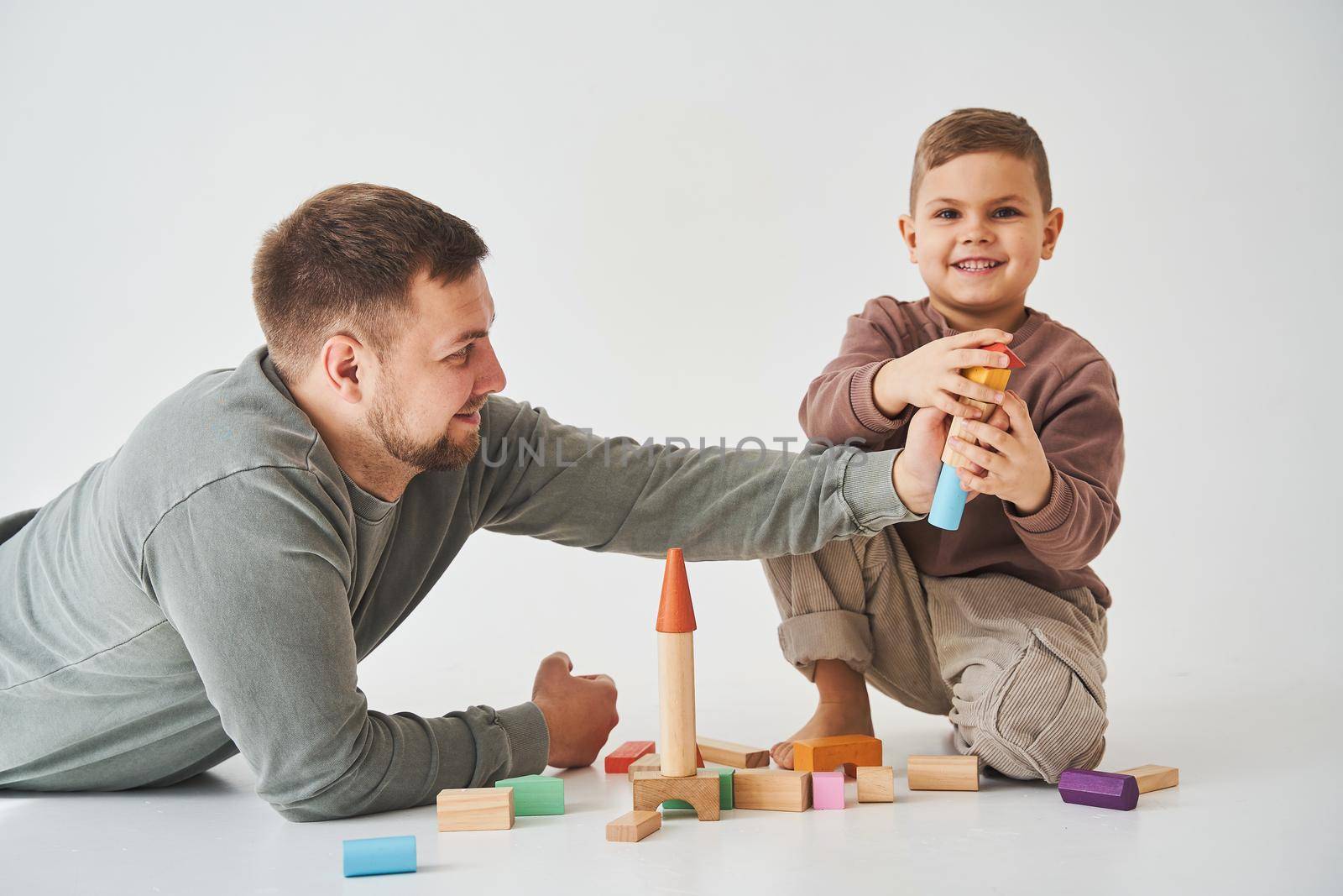 Caring dad plays with cheerful son with toy wooden cubes on white background. Fatherhood and child care