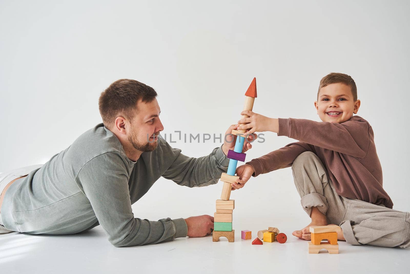 Caring dad helps his son to play on the floor on white background. Father and child build tower of colorful wooden bricks and have fun together. by Rabizo