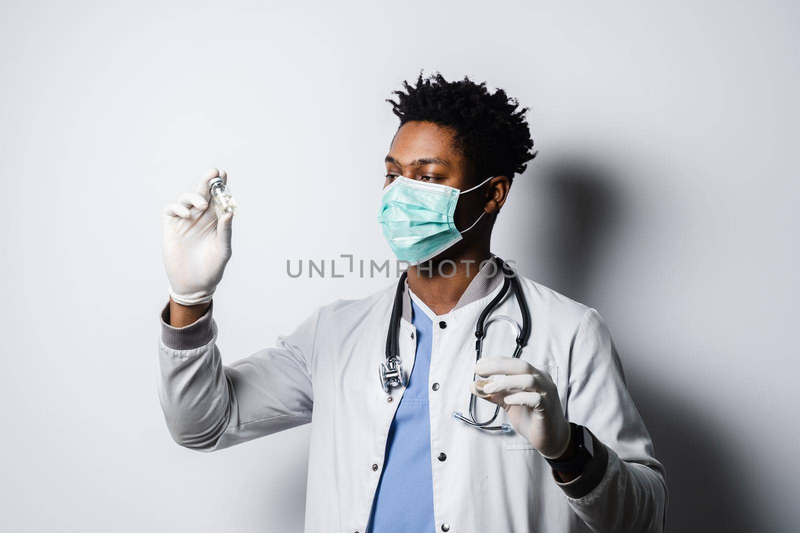 Black doctor in medical mask is holding vaccine on white background. Handsome African doctor with coronavirus covid-19 vaccine. by Rabizo