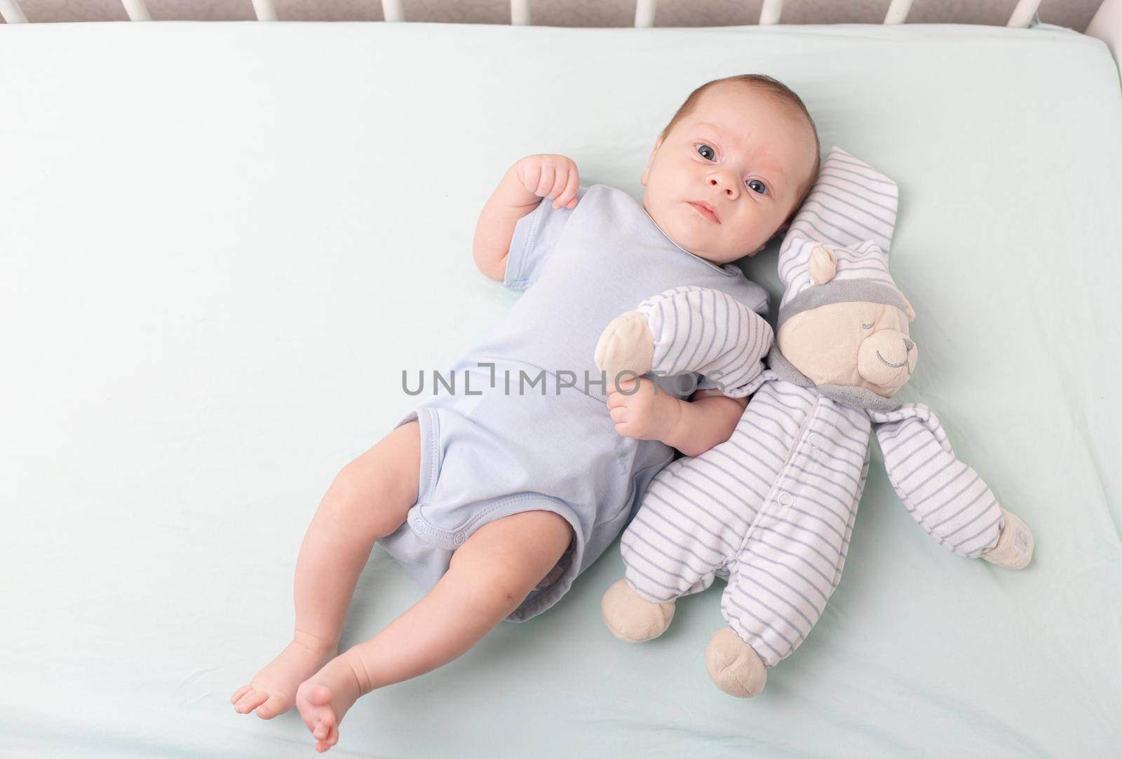 The baby is lying in a crib with a teddy bear . The baby is 0-3 months old. Calm kid. An article about toys for kids. A soft toy. Copy Space