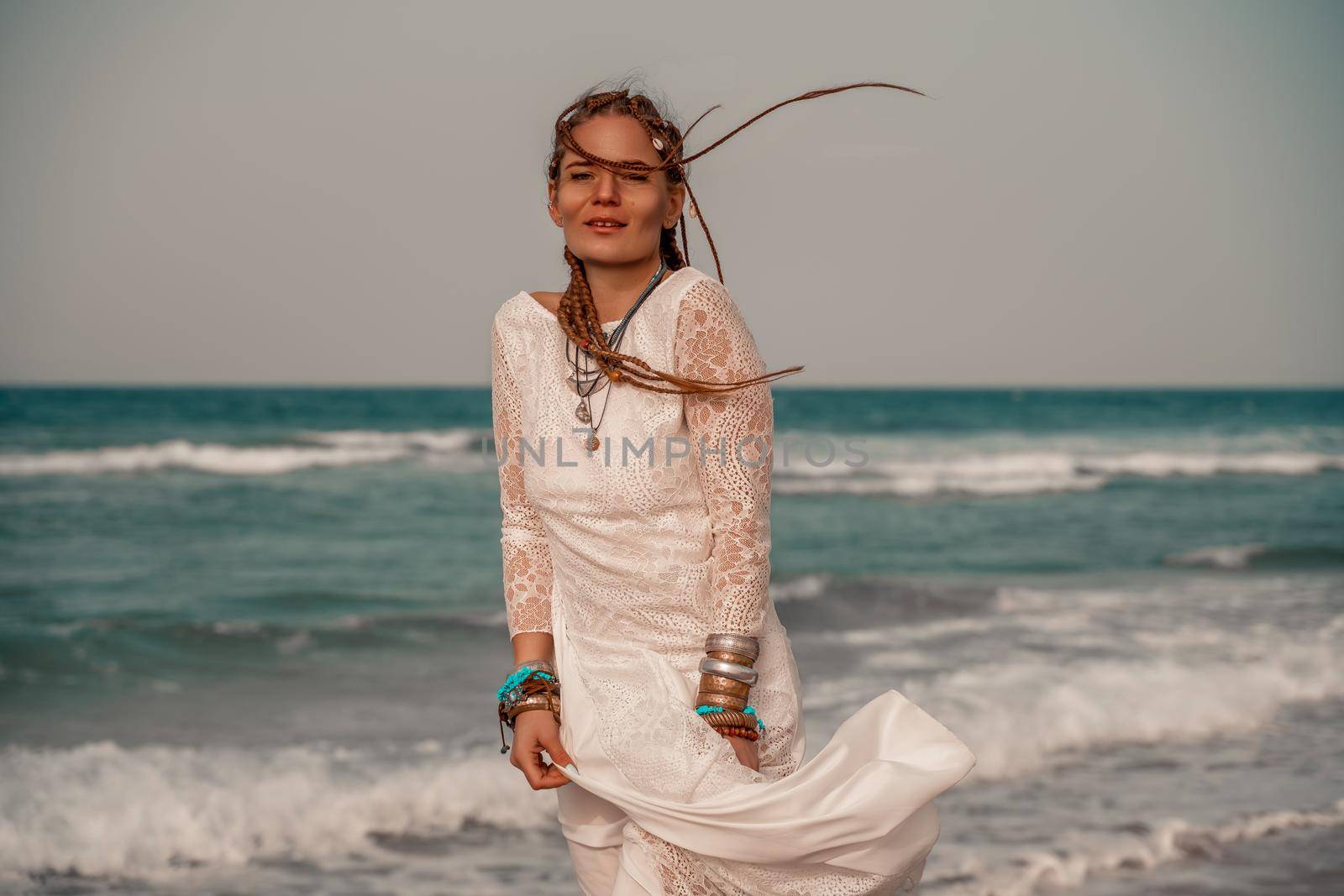 Model in boho style in a white long dress and silver jewelry on the beach. Her hair is braided, and there are many bracelets on her arms