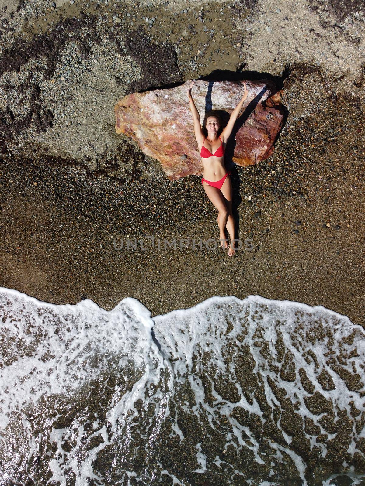 Top aerial view on a young sensual blonde woman in red bikini lying on the rock near water at sea and enjoys the sea waves. Dreams holidays and weekend vacation in summer time. by panophotograph