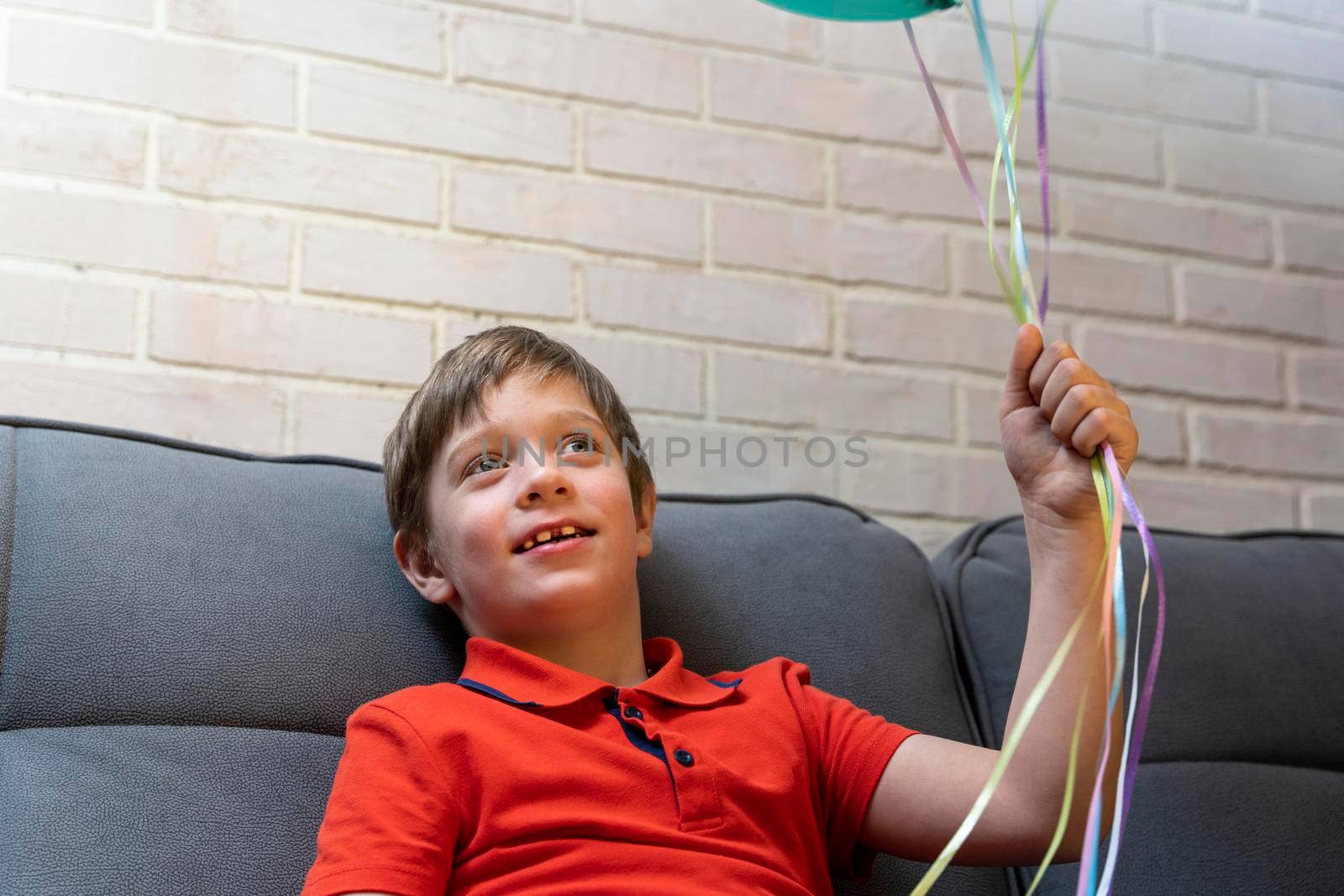 a happy European preschool boy is sitting on the sofa and smiling. The boy holds balloons inflated with helium by the ribbons. Birthday Celebration