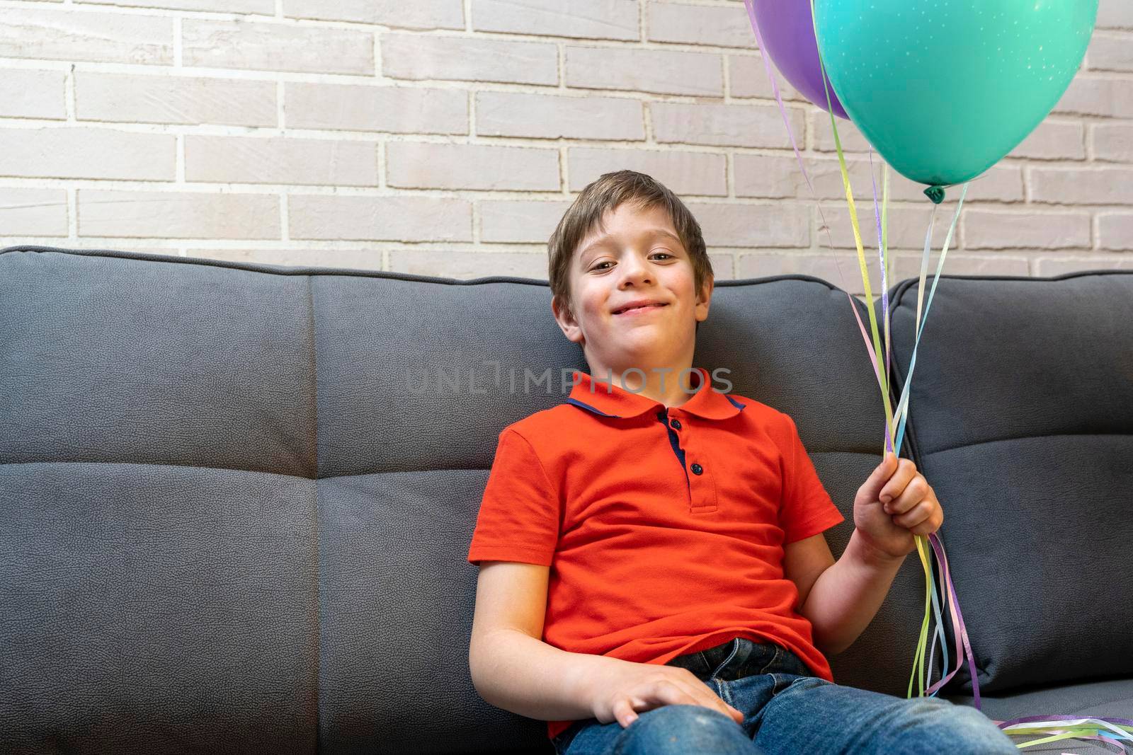 a happy European preschool boy is sitting on the sofa and smiling. The boy holds balloons inflated with helium by the ribbons. Birthday Celebration