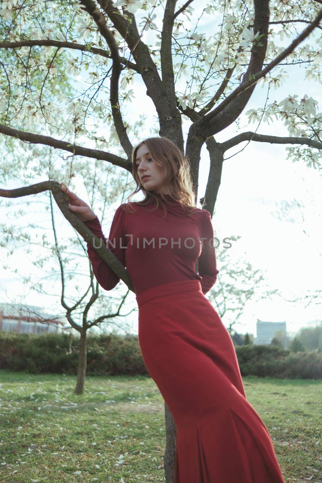A blonde girl in red leans on a tree in the park. the concept of unity with nature