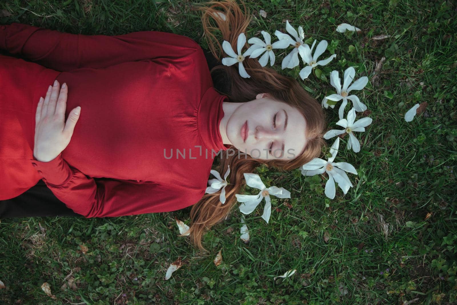 a blonde girl in red lies in the spring on the grass surrounded by magnolia flowers