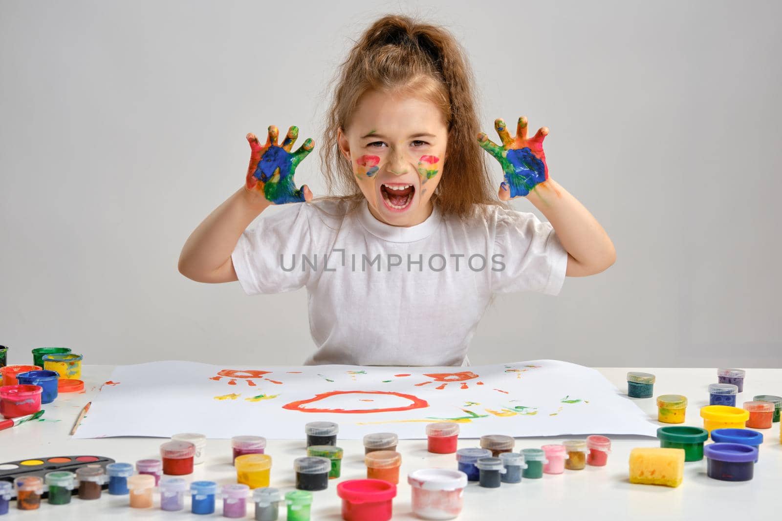 Pretty brunette child artist with a ponytail, in a white t-shirt, is sitting at a table with a whatman and a lot of multi-colored paints on it. She is posing with painted face and hands, scaring us. Art studio. Isolated on white background. Drawing process. Medium close-up.