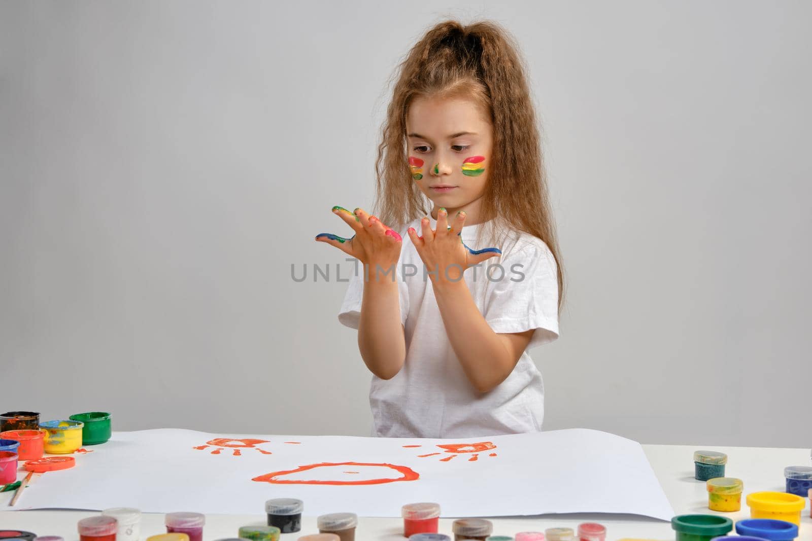 Little girl in white t-shirt sitting at table with whatman and colorful paints, painting on it with her hands. Isolated on white. Medium close-up. by nazarovsergey