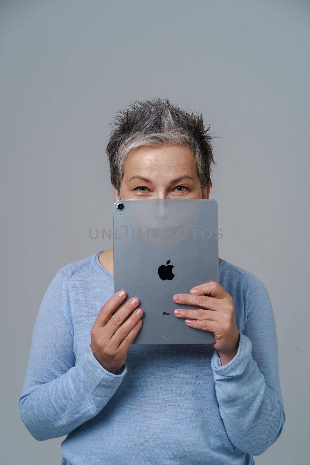 Mature grey haired woman hide hide behind apple ipad posting fake information or bad comments social media with drown evil face or alter ego on tablet isolated on white. Kyiv, Ukraine, May13, 2022 by LipikStockMedia