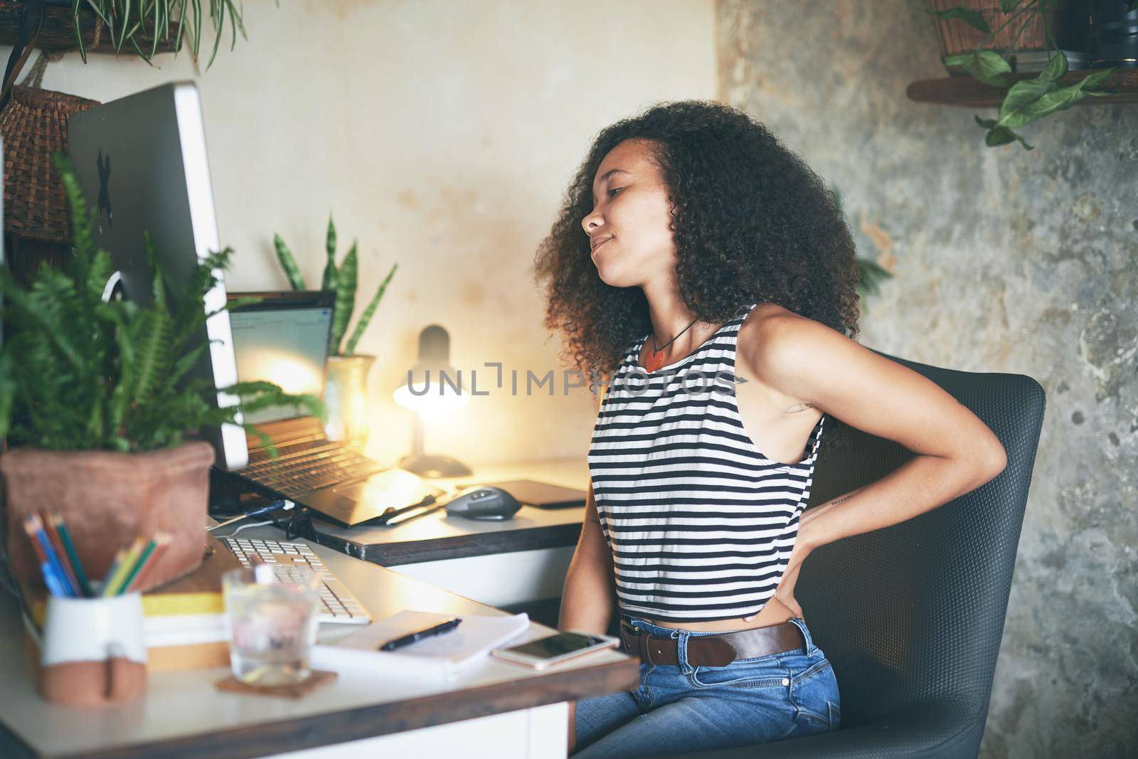 Shot of a young african woman sitting alone and suffering from back ache while using her computer to work from home stock photo