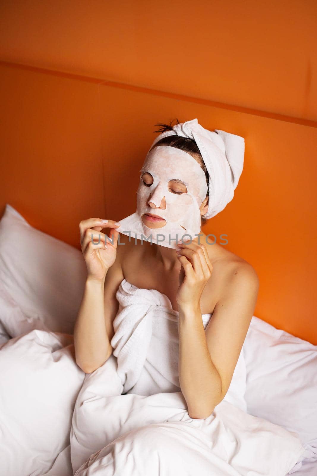 Young girl with clean skin with a mask on her face, sitting in bed, resting after a shower, relaxing at home. Close-up