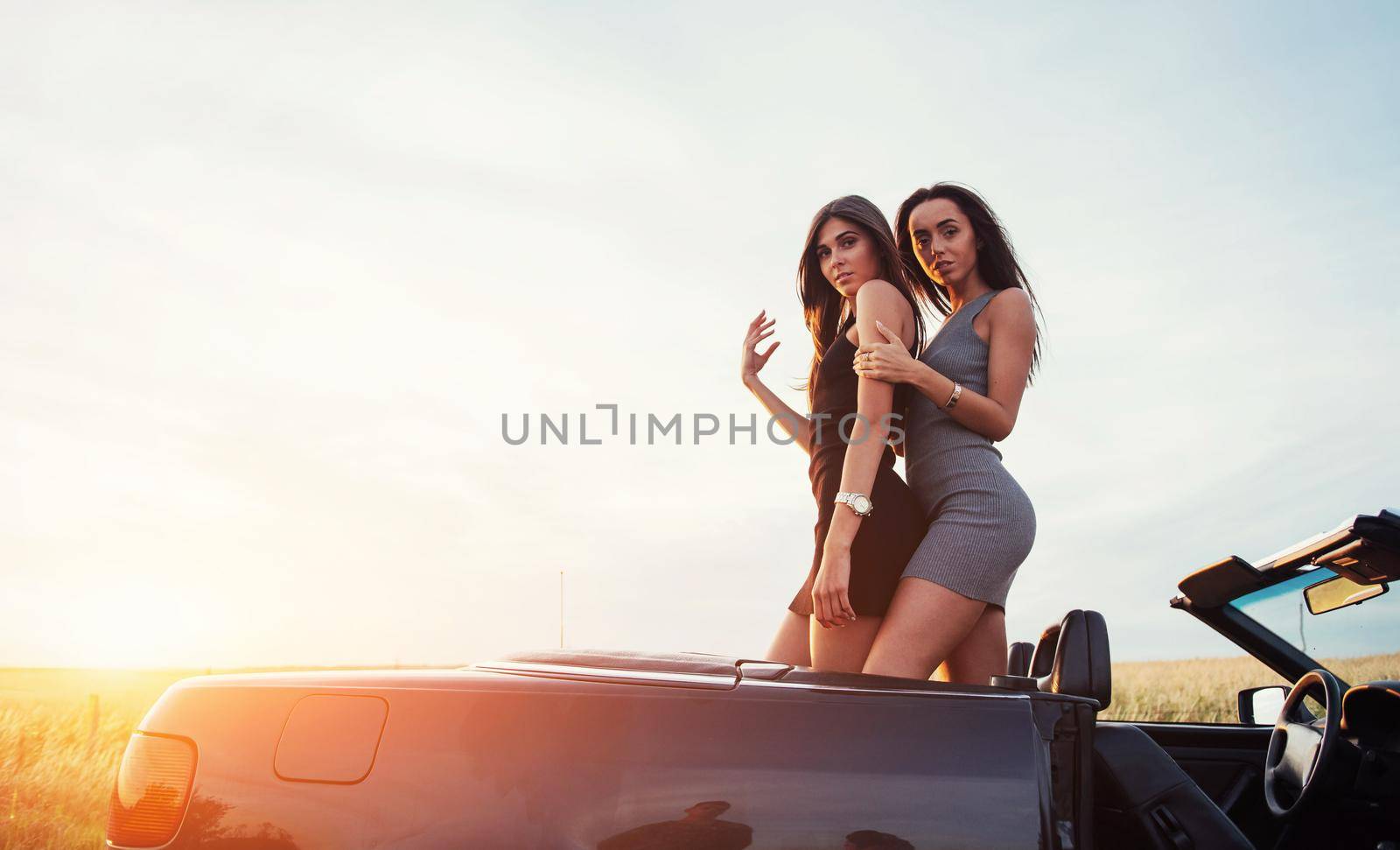 Two girls happy to pose next a black car against the sky on a fantastic sunset.