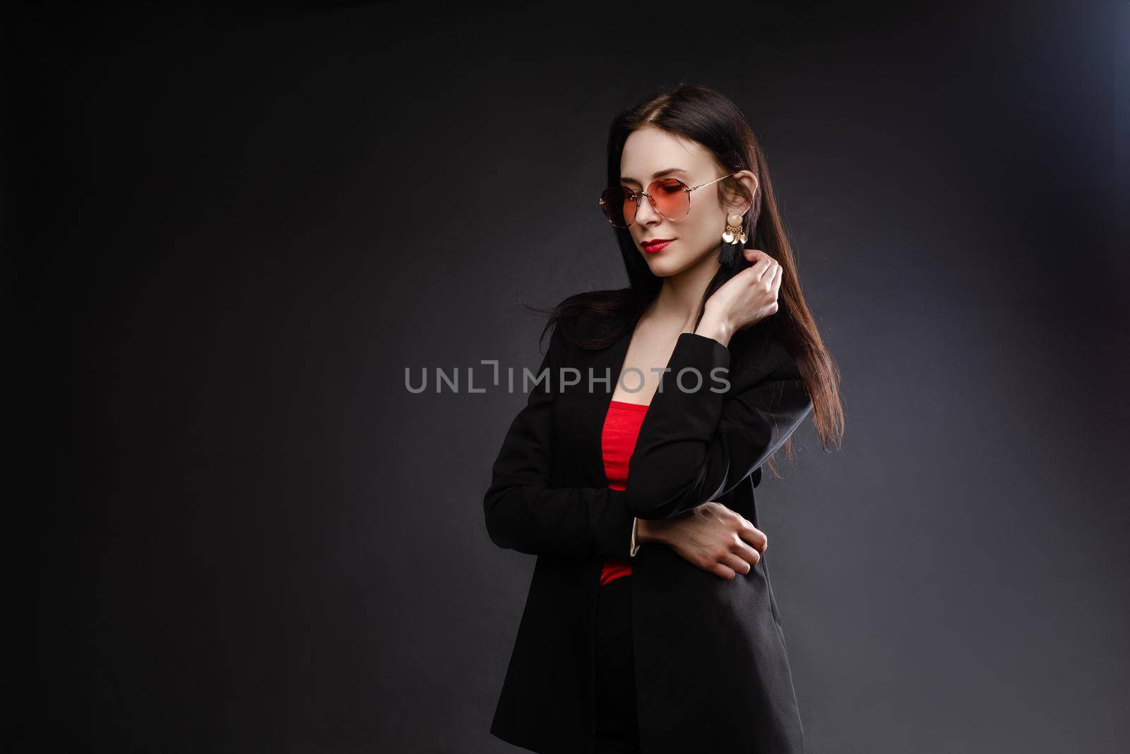 Portrait of young beautiful woman thoughtfully looking down. Brunette model in elegant costume posing at camera . Pretty lady in red sunglasses leaning one hand on other and holding it near face.