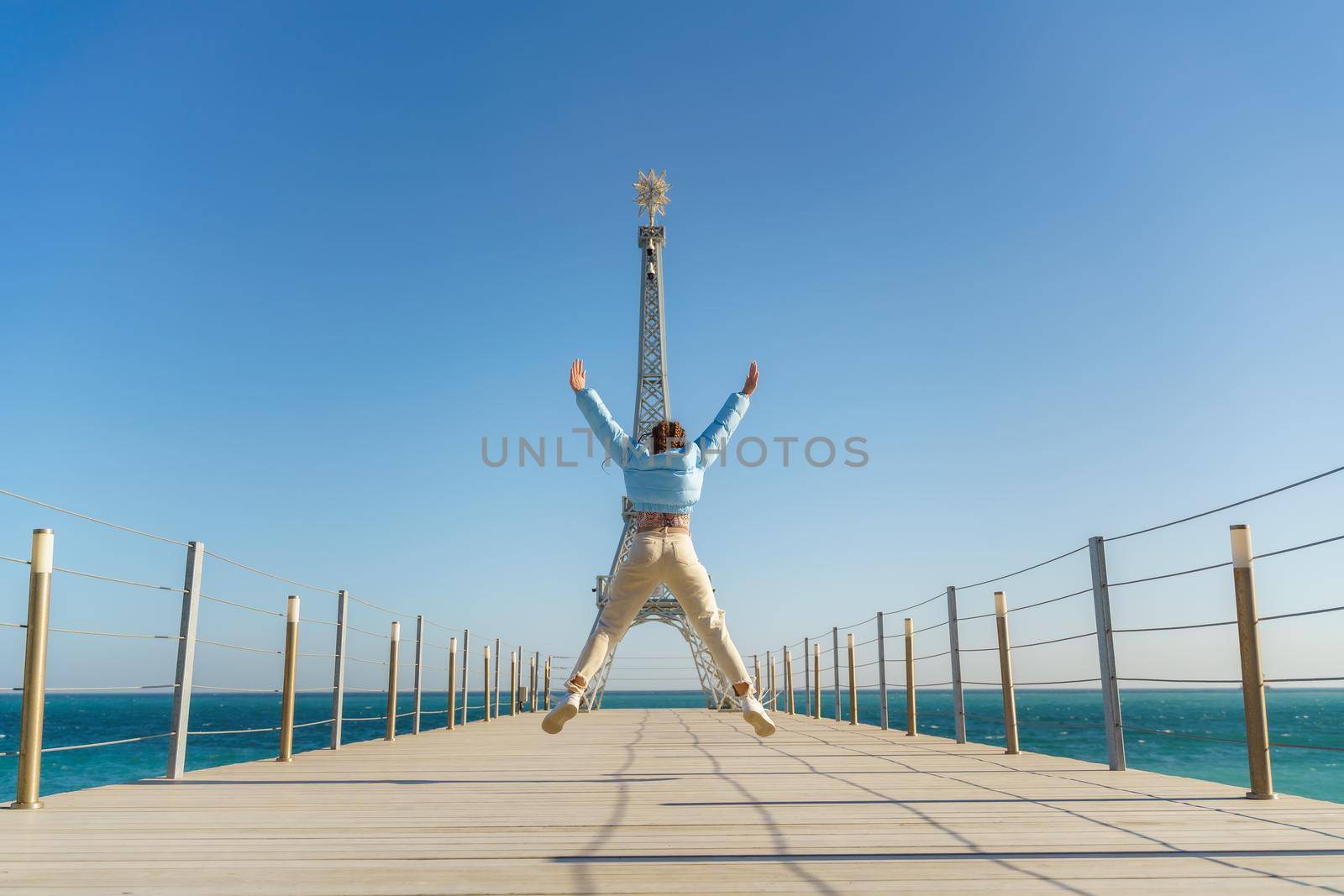 Large model of the Eiffel Tower on the beach. A woman walks along the pier towards the tower, wearing a blue jacket and white jeans. by Matiunina