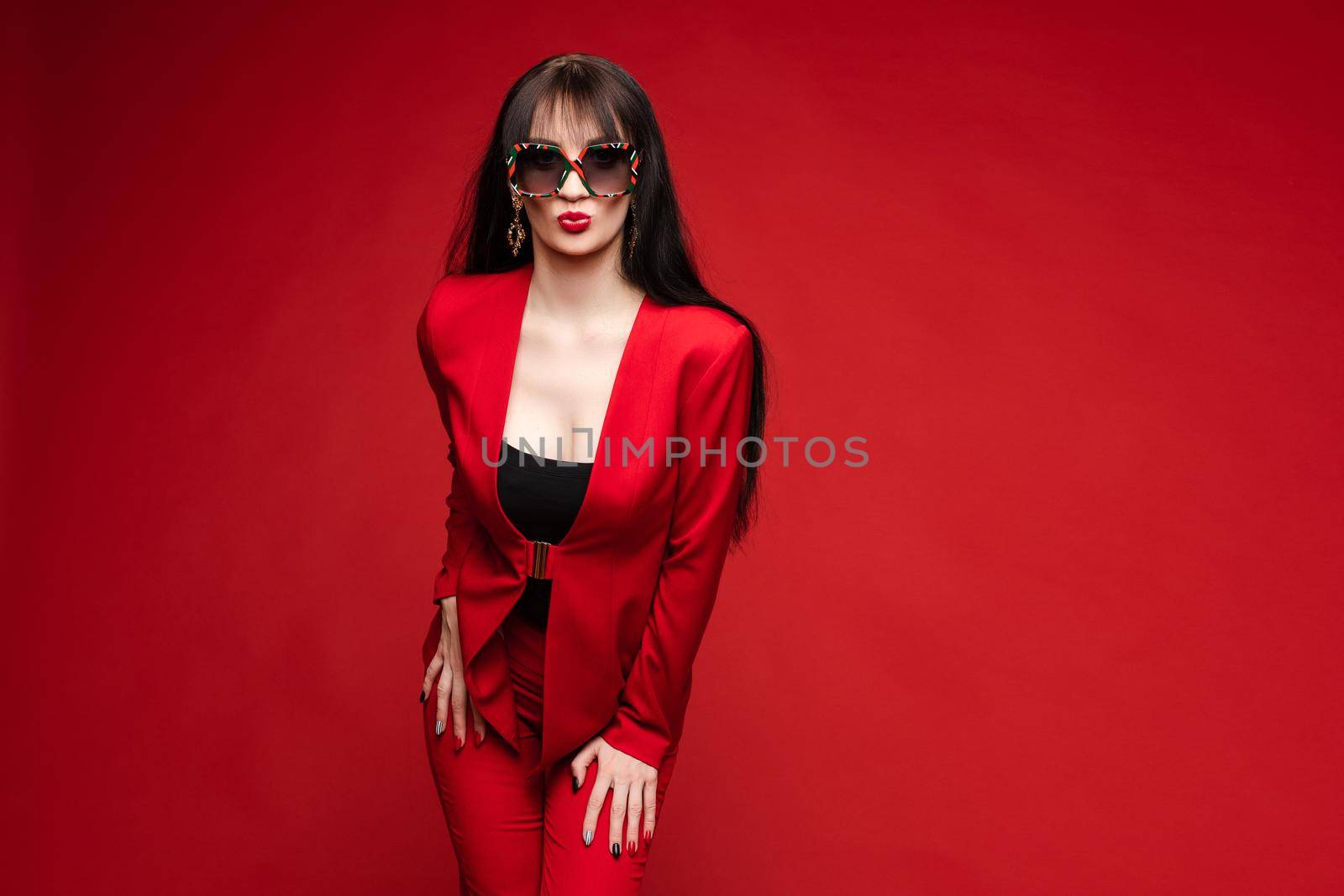 Side view of glamorous brunette posing in red smart suit and heels on red isolated background in studio. Young woman keeping glasses in hand and looking down. Concept of beauty and fashion.