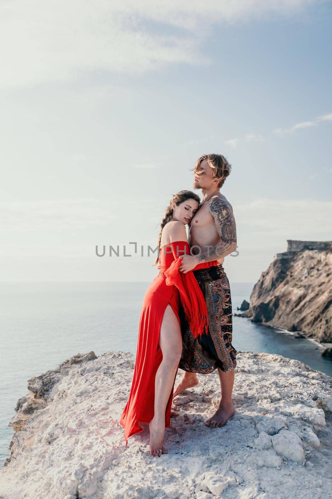 Portrait of tattooed man and sensual young brunette woman in red dress, happily dancing outdoors isolated on blurry sea background with natural bokeh in soft warm sunset backlight. Selective focus