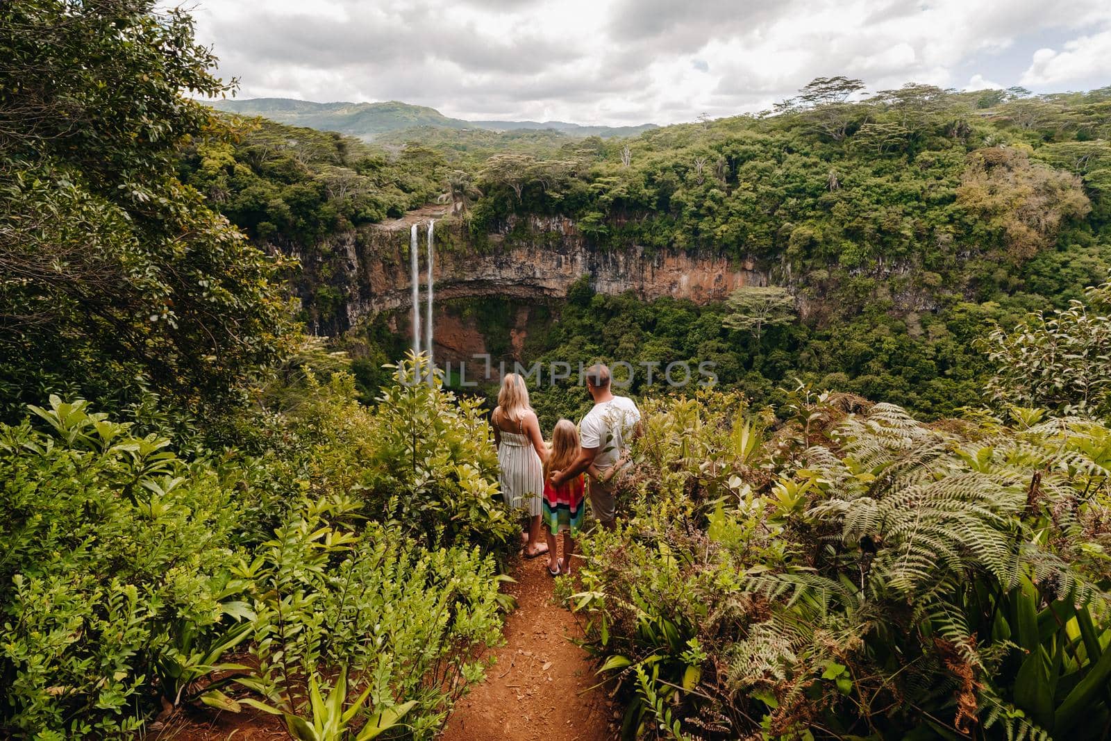 a family, a man, a woman and a daughter, stand on the edge of a cliff near a waterfall in Chamarel Park on the island of Mauritius.A couple with their daughter in the jungle of the island of Mauritius looks at a large waterfall falling down