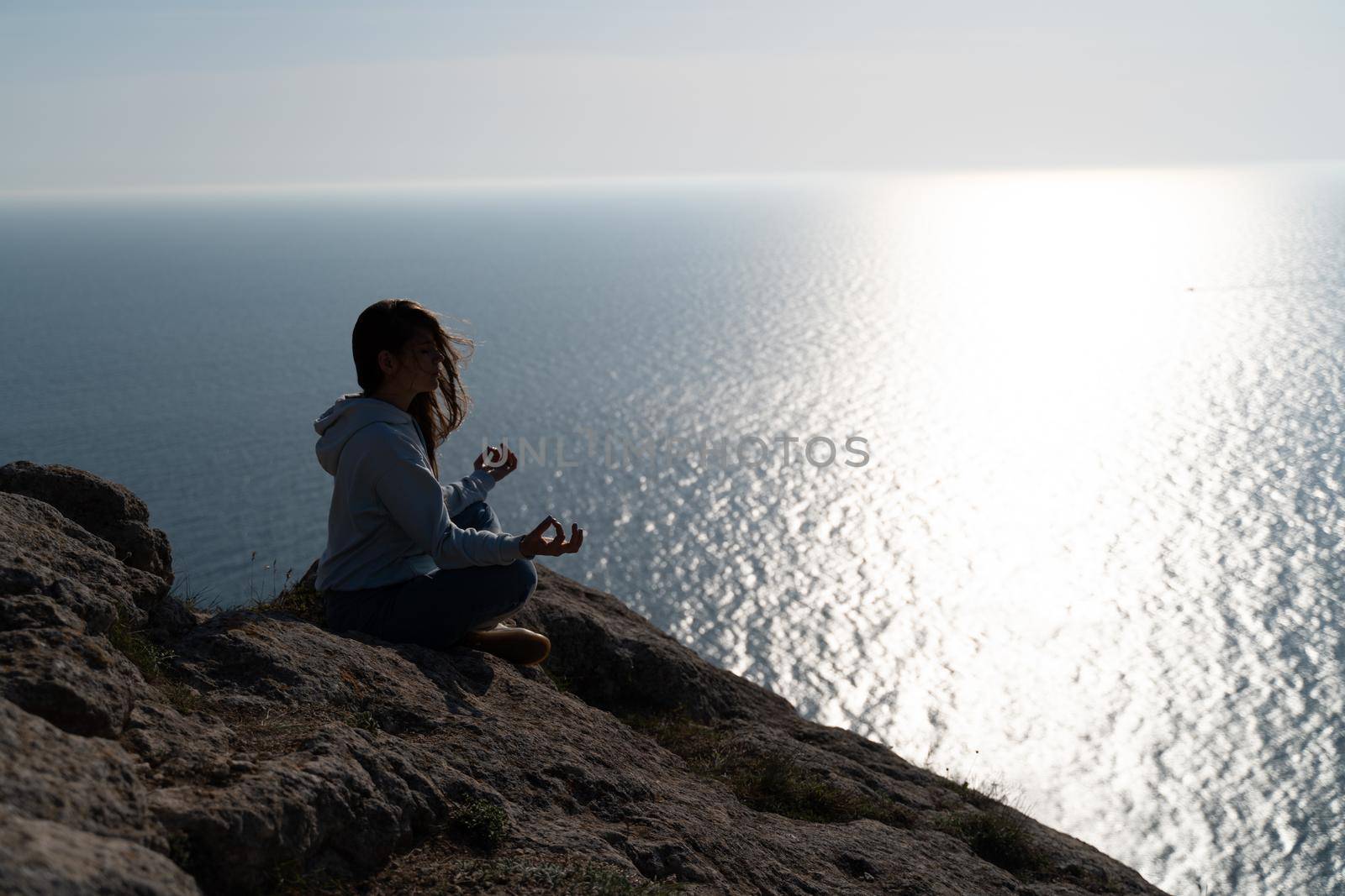 Woman tourist enjoying the sunset over the sea mountain landscape. Sits outdoors on a rock above the sea. She is wearing jeans and a blue hoodie. Healthy lifestyle, harmony and meditation by Matiunina