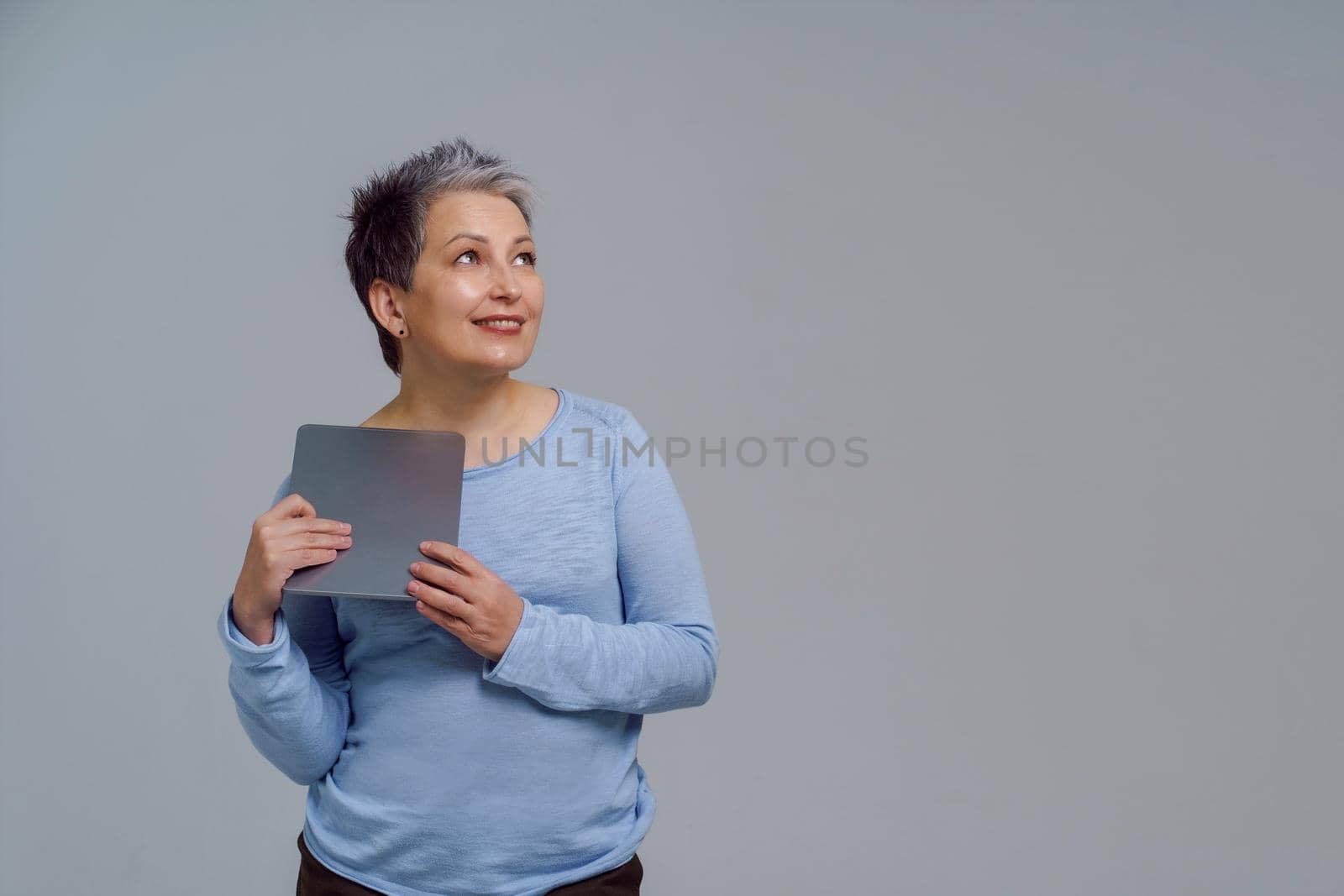 Dreaming beautiful mature grey haired woman in 50s holding digital tablet working or shopping online or checking on social media. Pretty woman in blue blouse isolated on white background. Toned image.