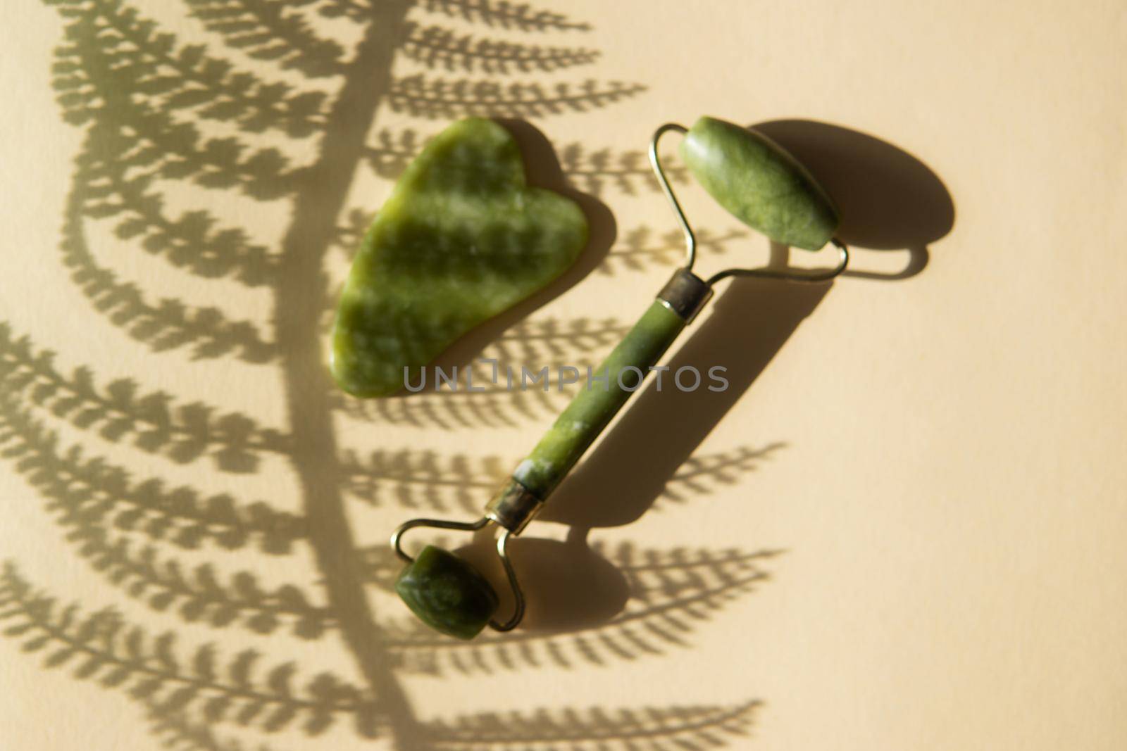 Jade Gua sha scraper and face roller massager on a cork round stand with a monstera leaf. Hard light, shadows, the concept self-care. Facial care. Zero waste. Lifting and toning treatment at home.