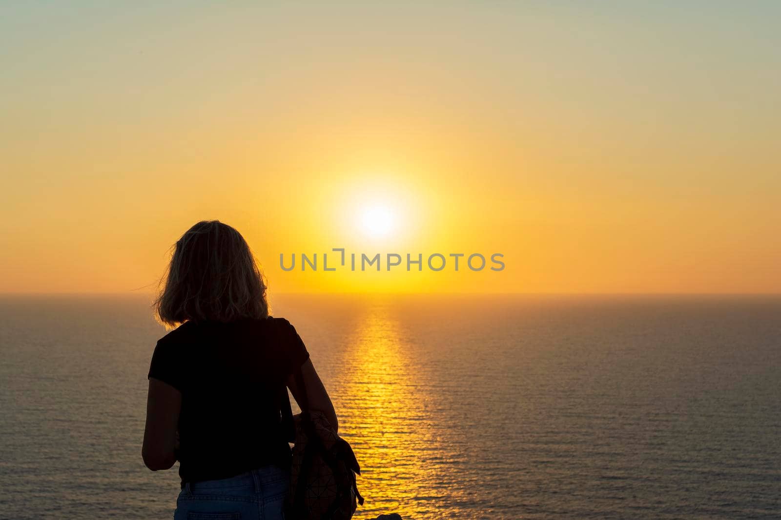 Silhouette of a woman at orange sunset on a cliff overlooking the sea at Palaiokastro castle of ancient Pylos. Greece.