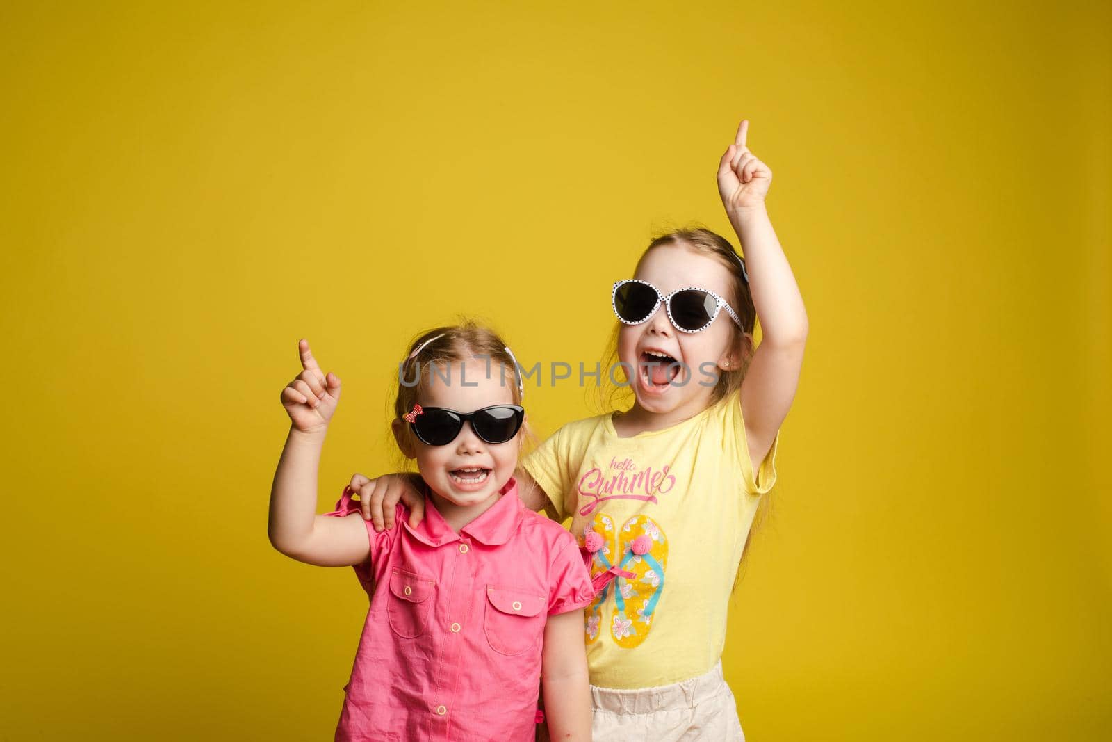 Two happy beautiful stylish little girl wearing sunglasses posing isolated at yellow studio background medium shot. Adorable laughing female child friend enjoying friendship having positive emotion